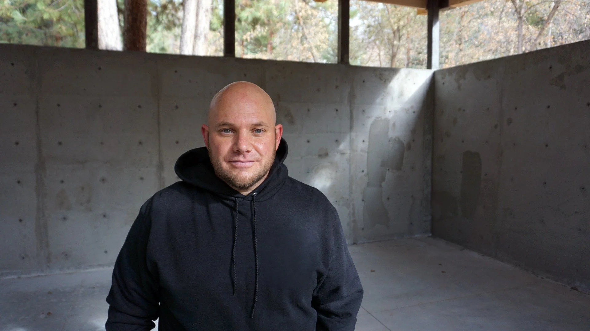 Black and white portrait of a bald man wearing a zipped-up hoodie, smiling slightly, against a plain white background.