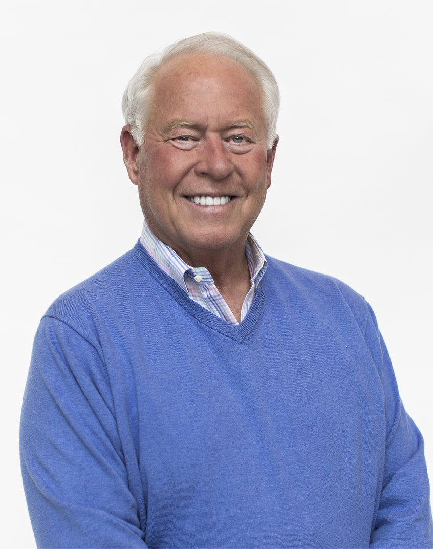 Portrait of an elderly man smiling, wearing a blue sweater over a collared checkered shirt, against a plain white background.
