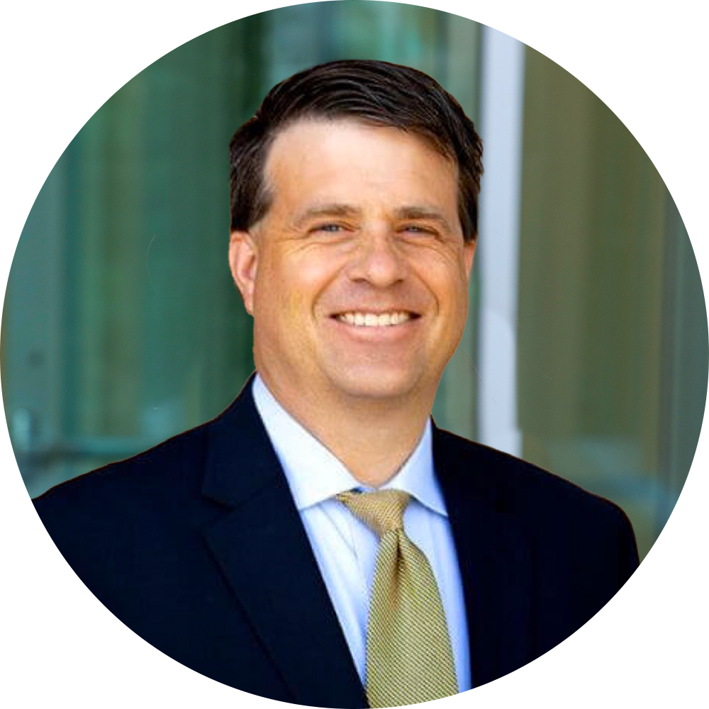 Headshot of a smiling man with brown hair, wearing a dark suit, light blue shirt, and yellow tie, standing outdoors with a glass building in the background.