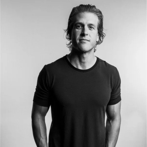 Black and white photo of a young man with long hair, wearing a dark T-shirt, looking at the camera with a neutral expression.