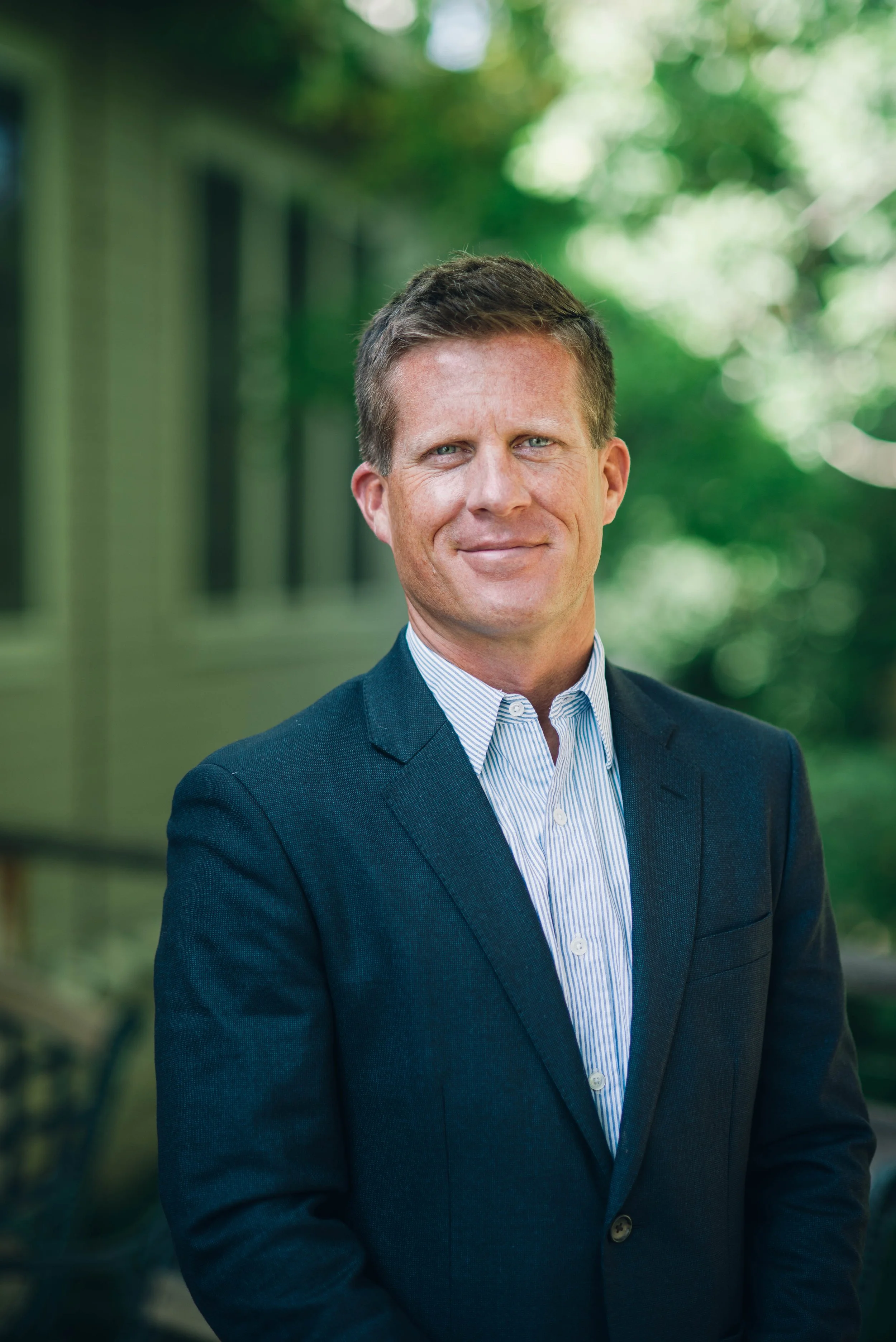 A black and white portrait of a man with short hair, wearing a collared shirt, outdoors with foliage in the background.