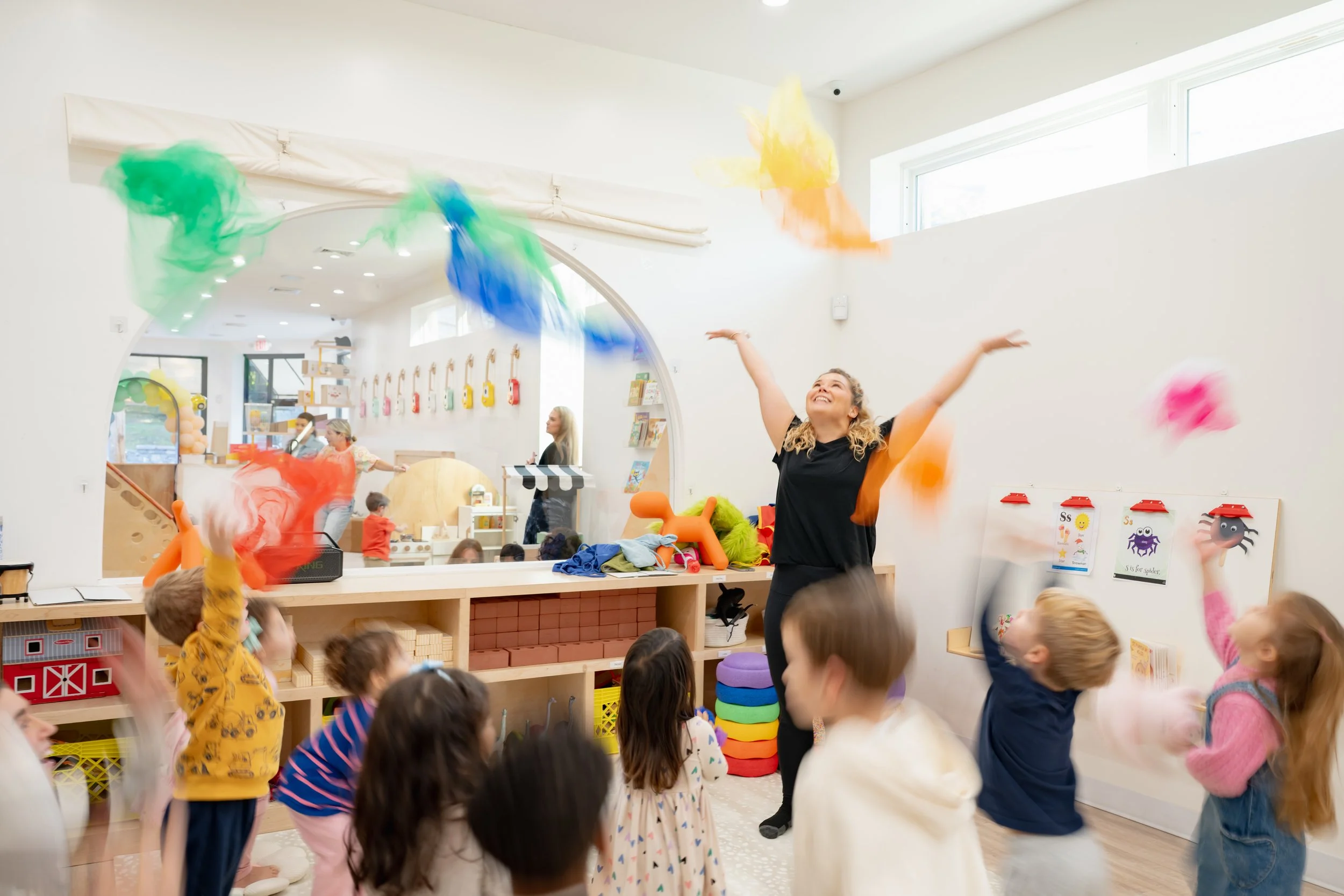 A teacher with a headset leading a small group of children seated on circular cushions in a colorful classroom.
