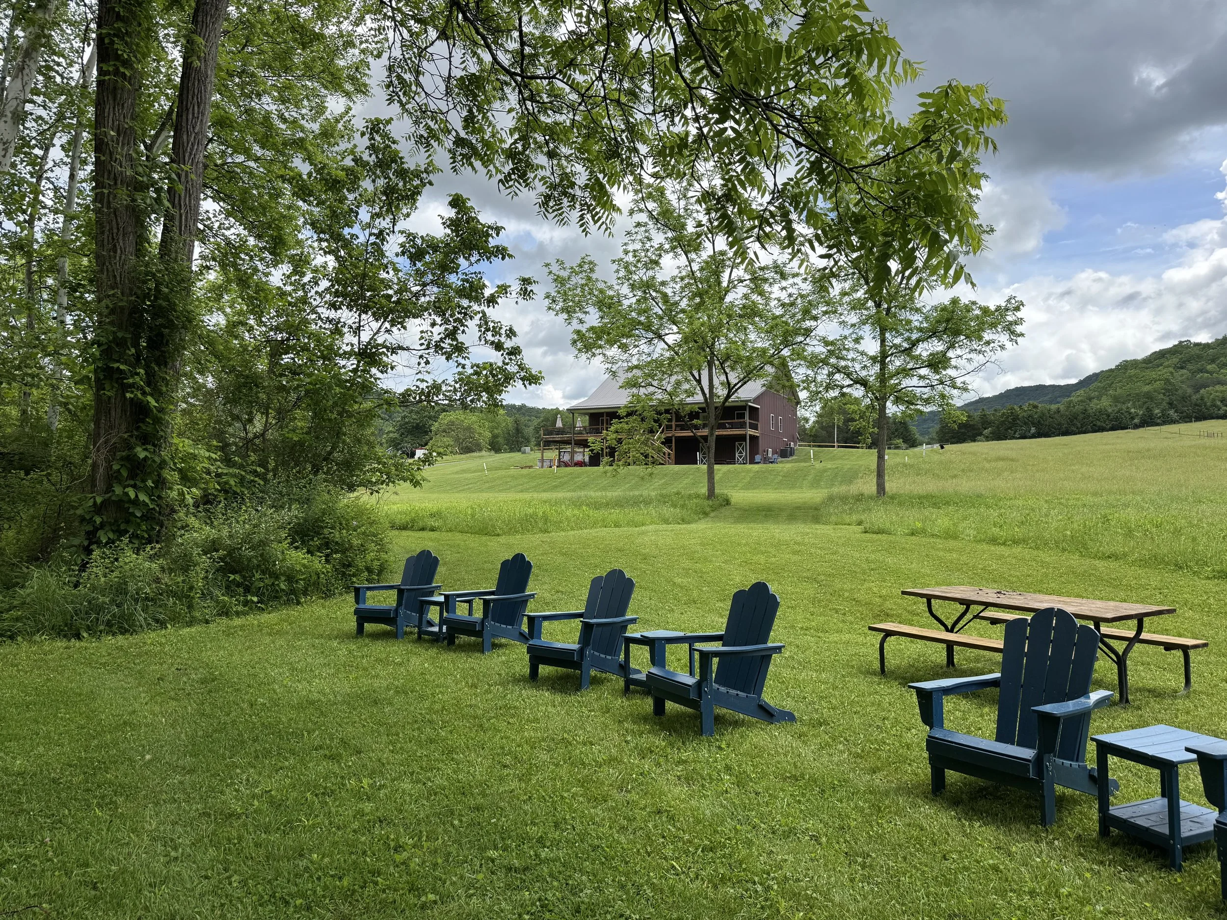 Adirondack chairs beside Indian Creek offer a peaceful place to relax at Old World Libations, with open fields, wooded hills, and the winery tasting room just up the slope. It is a quiet Appalachian setting where guests can enjoy wine, mead, cider, a