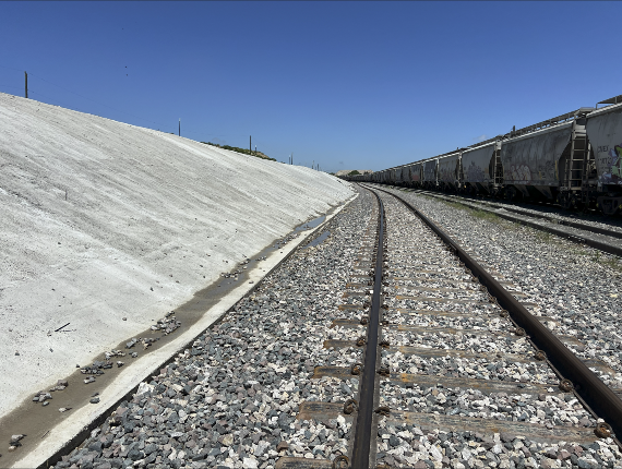Railroad tracks curving beside a line of freight train cars and a concrete-lined embankment under a clear blue sky