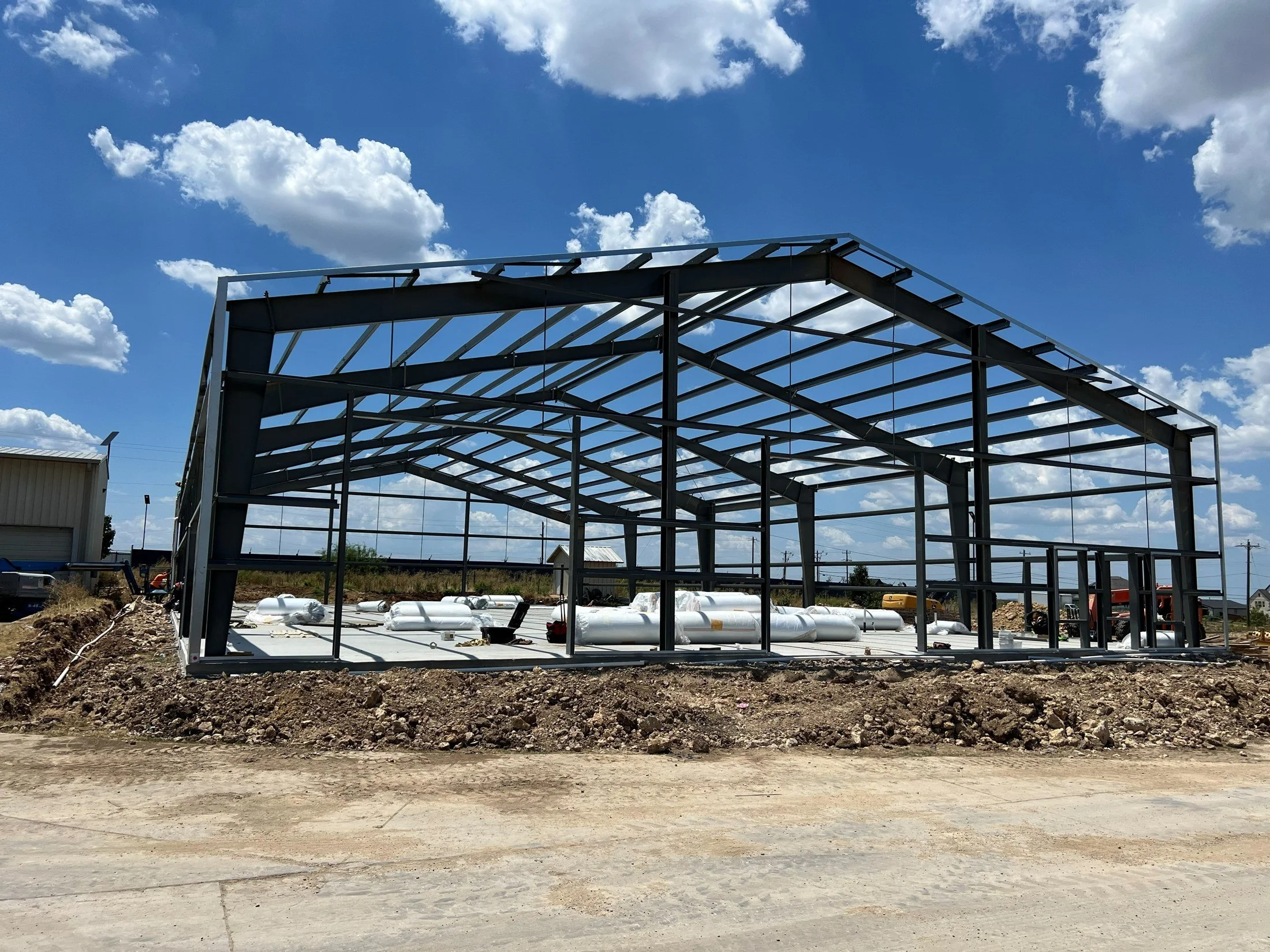 Steel frame of a commercial building under construction on a concrete foundation beneath a blue, cloud-filled sky