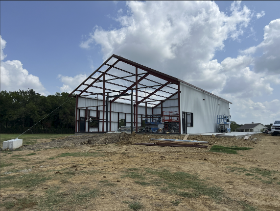 Large metal warehouse under construction with exposed steel framing, corrugated siding, and a dirt foreground beneath a partly cloudy sky