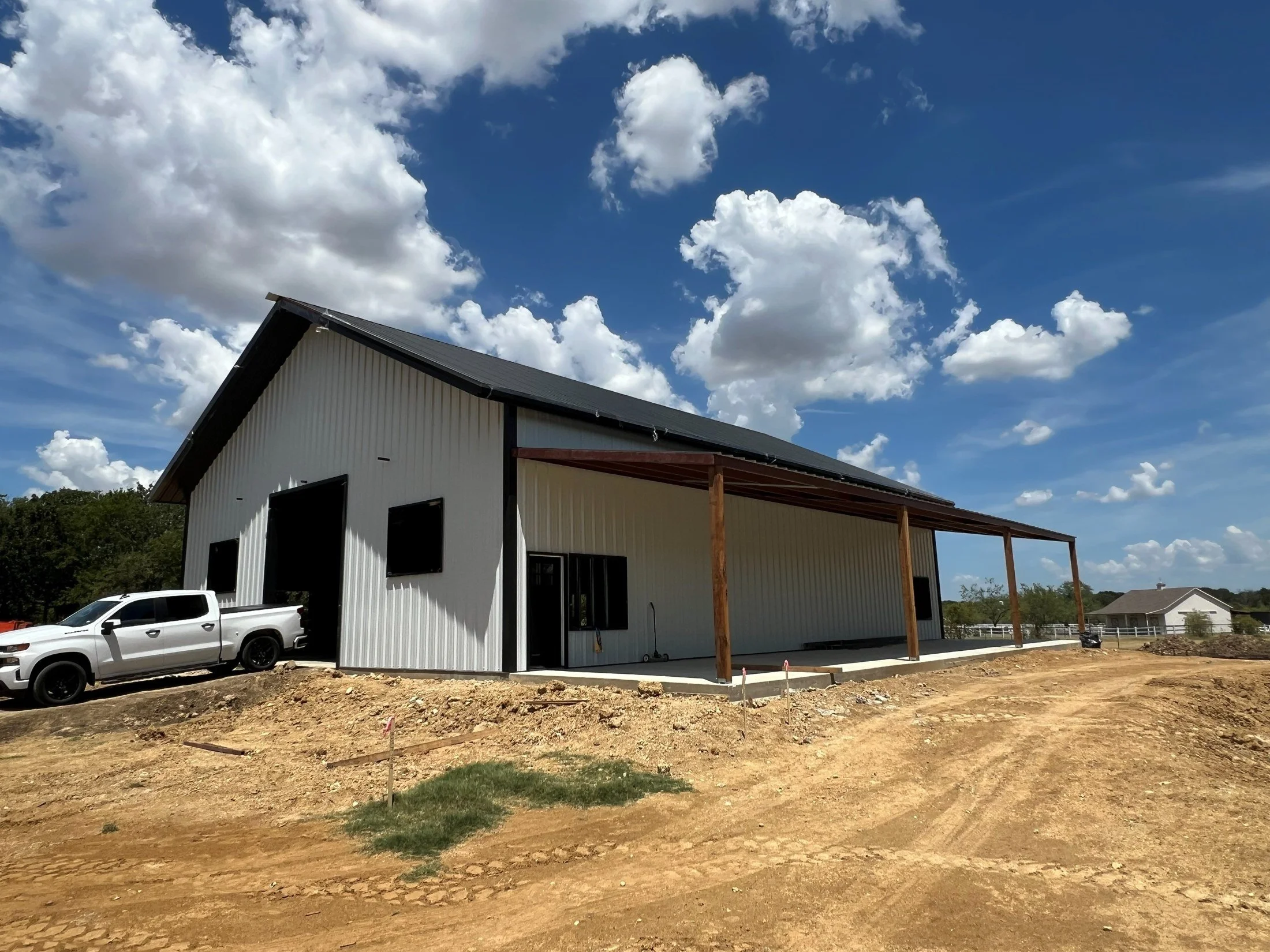 New white metal building under construction with a covered porch, white pickup truck, and bright clouds overhead