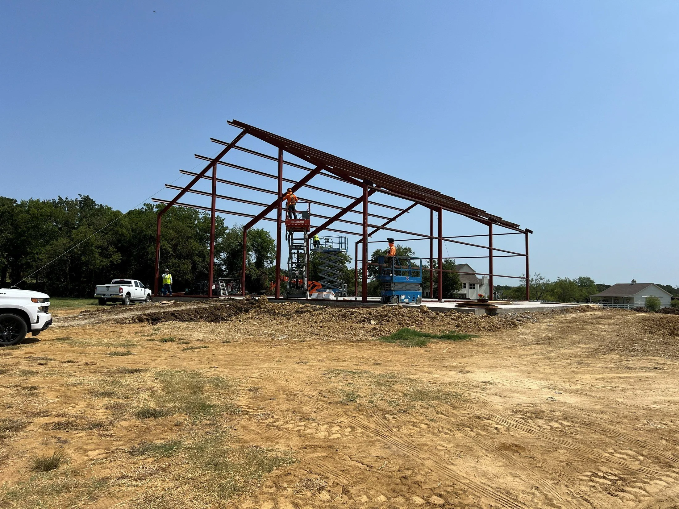 Construction workers assemble a steel-framed building using aerial lifts at a dirt construction site.