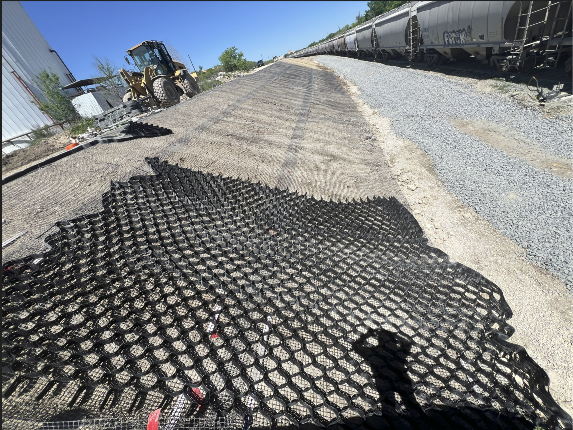 Black geogrid reinforcement mesh spread across a gravel rail yard, with a bulldozer and freight train cars in the background.