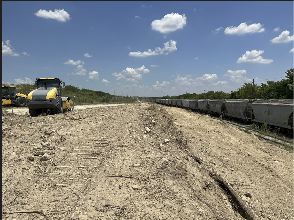 Construction site with road roller, dirt track, and freight train under a blue sky