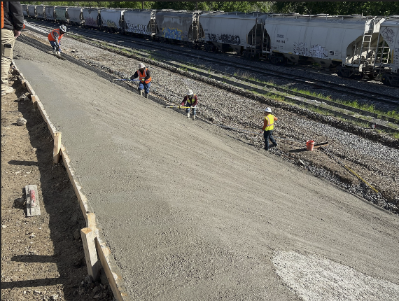 Construction workers paving a concrete path beside freight train tracks