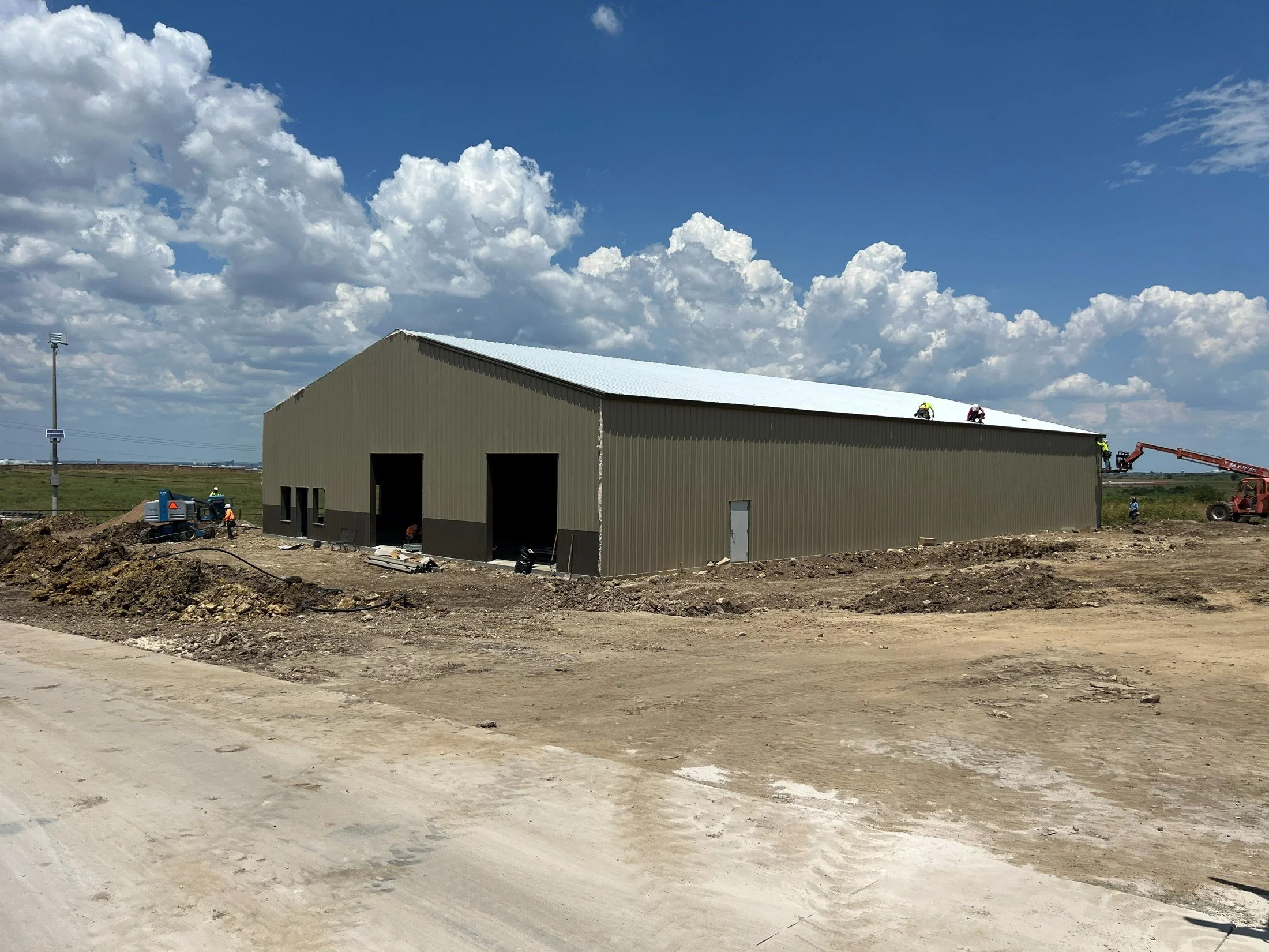 Construction workers and heavy equipment at a large metal warehouse under a bright blue sky with clouds