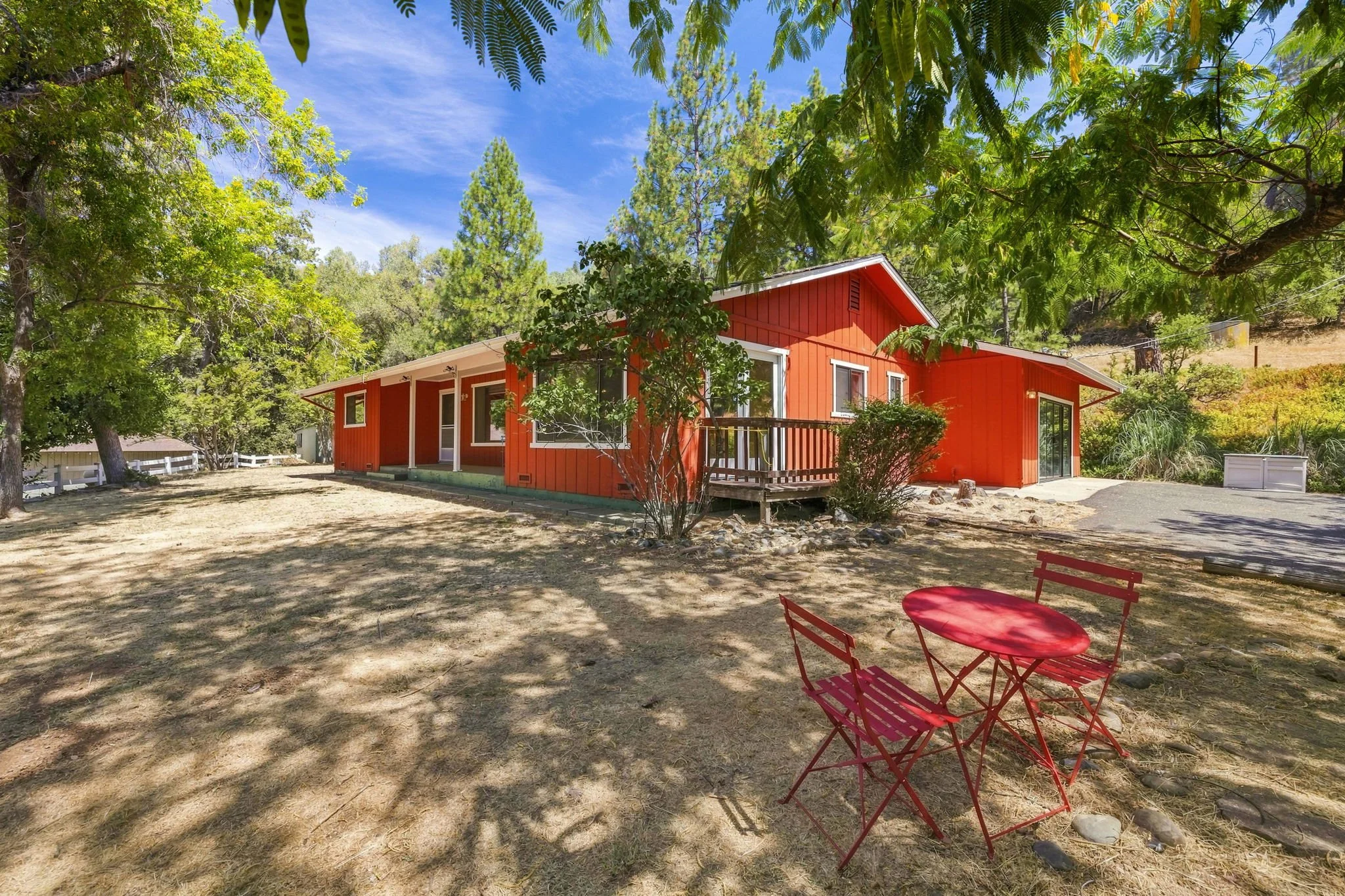 A red house sits in the shade under oak trees with a small red patio seating set in the foreground.