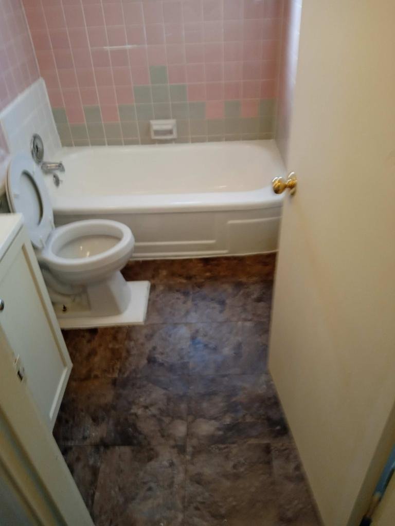 Bathroom with a bathtub, toilet, and tiled wall in shades of pink and green, with marble-patterned flooring and a closed door.