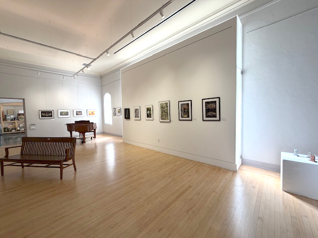 An art gallery with framed paintings on white walls, a wooden bench in the foreground, and a piano in the background.
