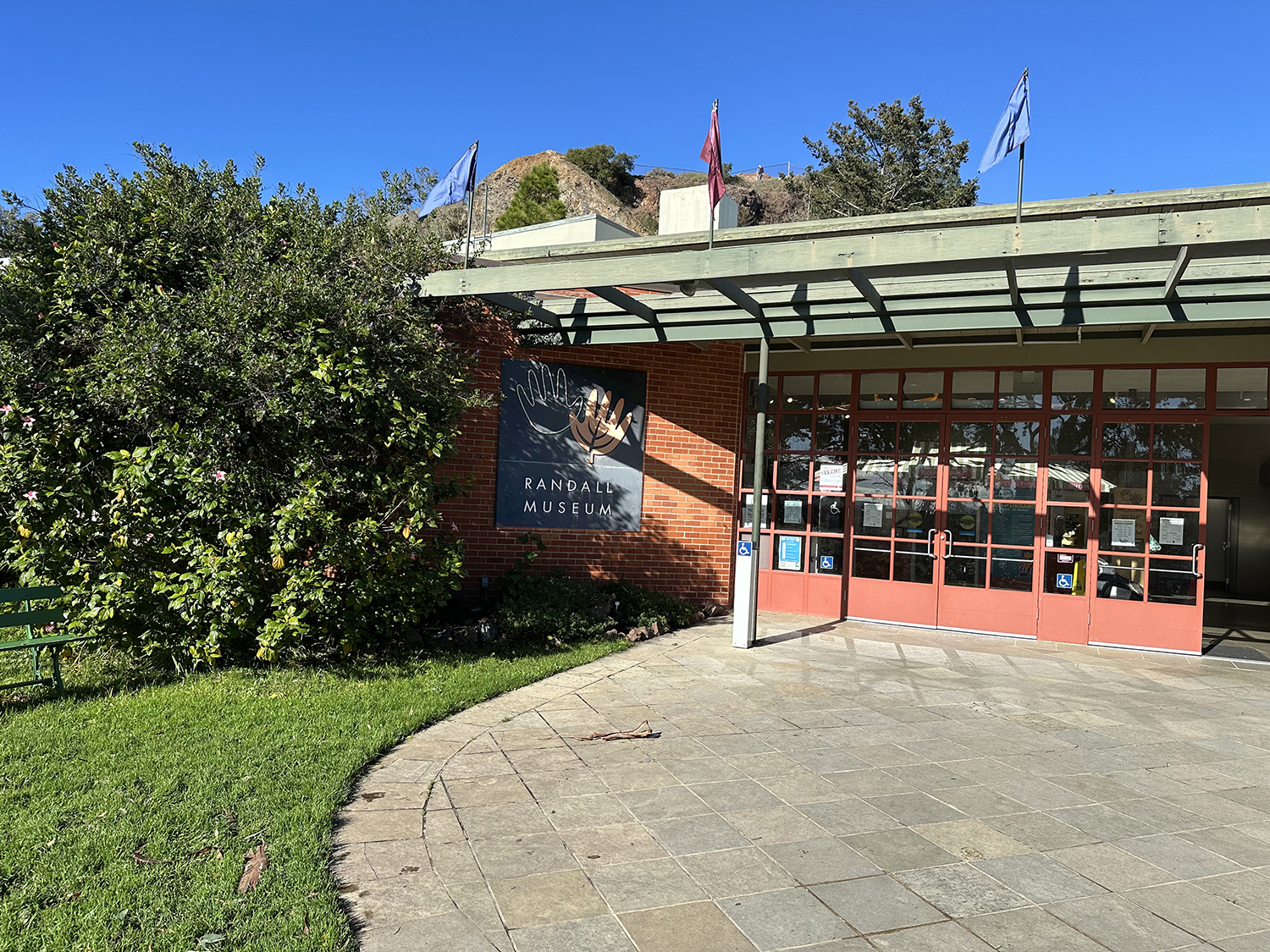 Exterior of the Randall Museum with brick walls, large glass doors, and a sign with two hands reaching towards each other. There are bushes and a green bench outside, and three flags flying on the roof.