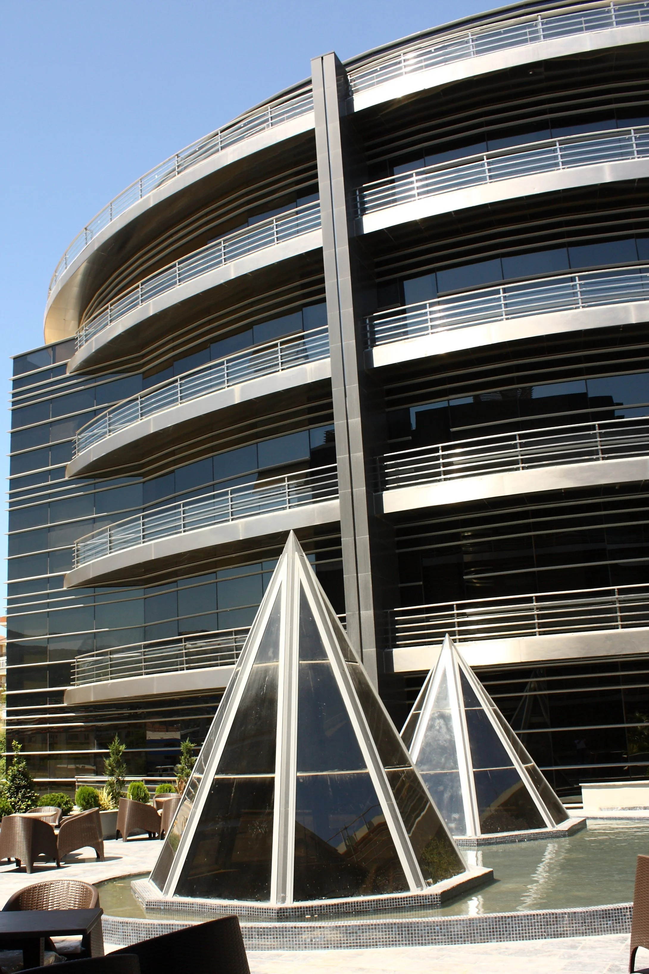 Modern building with curved balconies and glass windows, with pyramid-shaped glass structures and outdoor seating in the foreground.