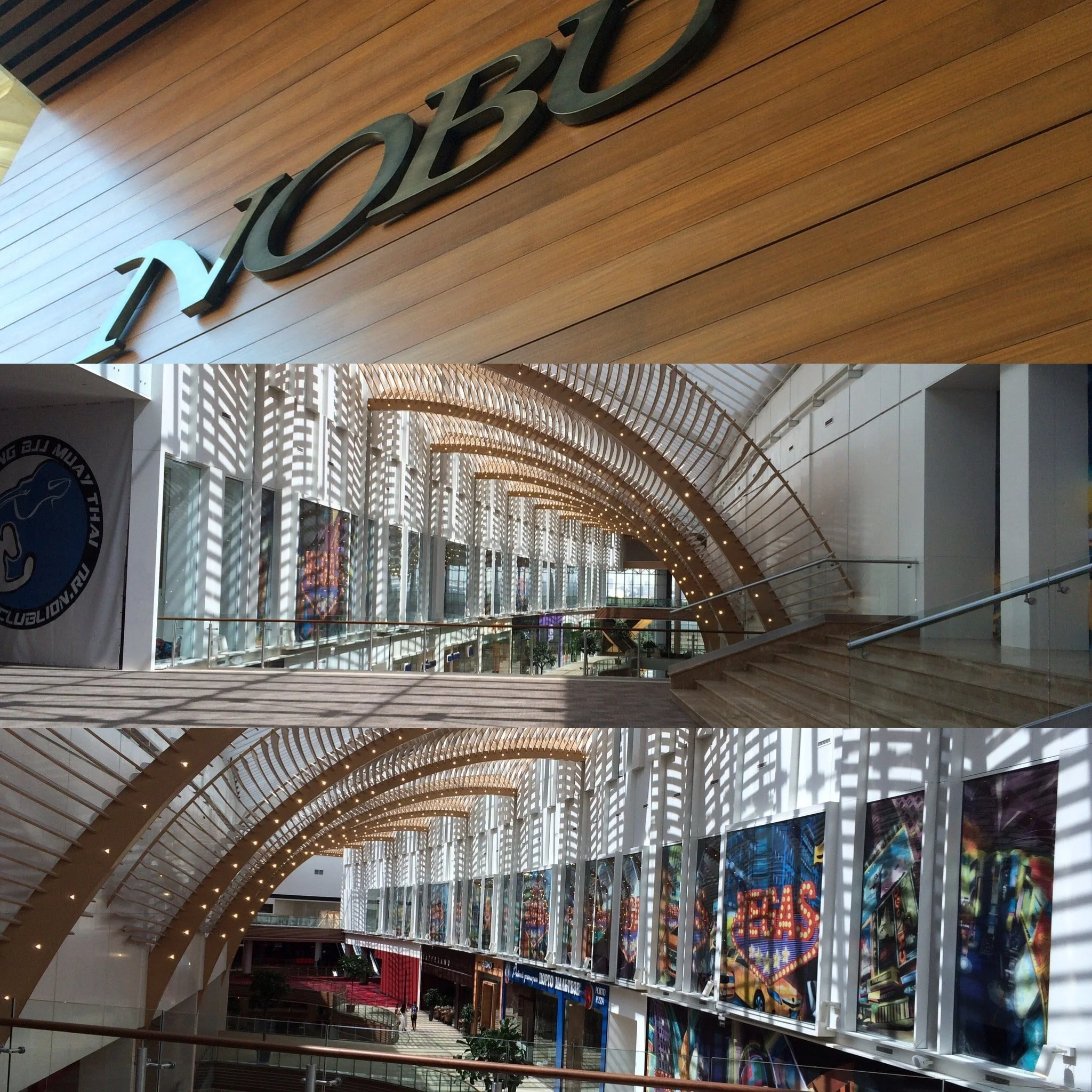 Interior view of a modern shopping mall with natural sunlight casting shadows through architectural details, displaying an arched ceiling, art displays, and a spacious walkway.