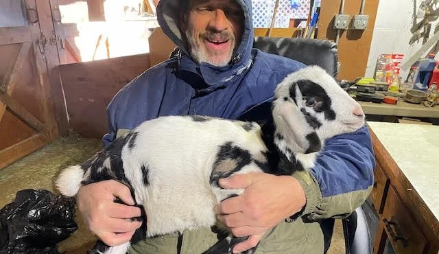 A smiling man holding a young black and white baby goat in a workshop or barn setting.