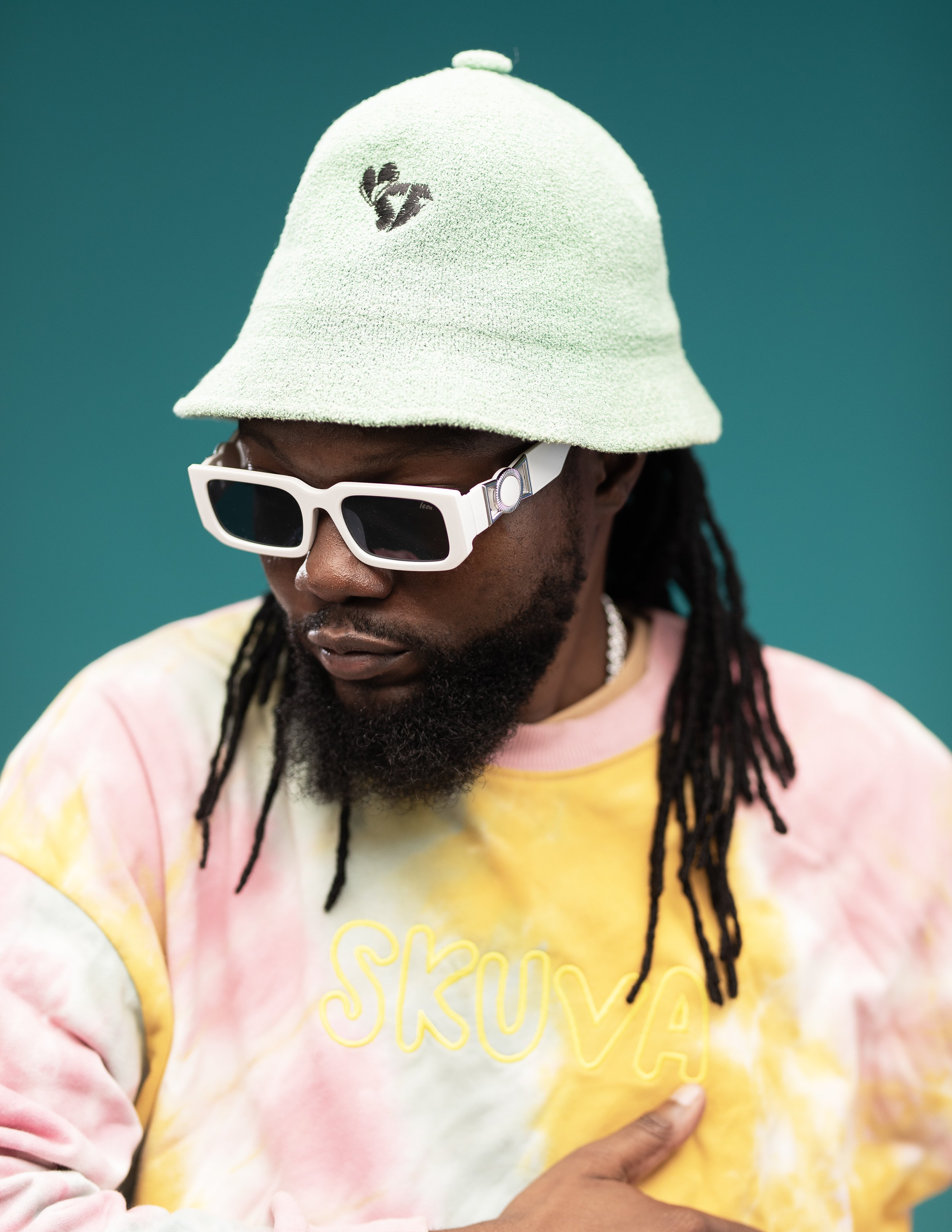 Person wearing a mint green bucket hat with a small logo, white sunglasses, and a pastel tie-dye sweatshirt with "SKUVA" text.