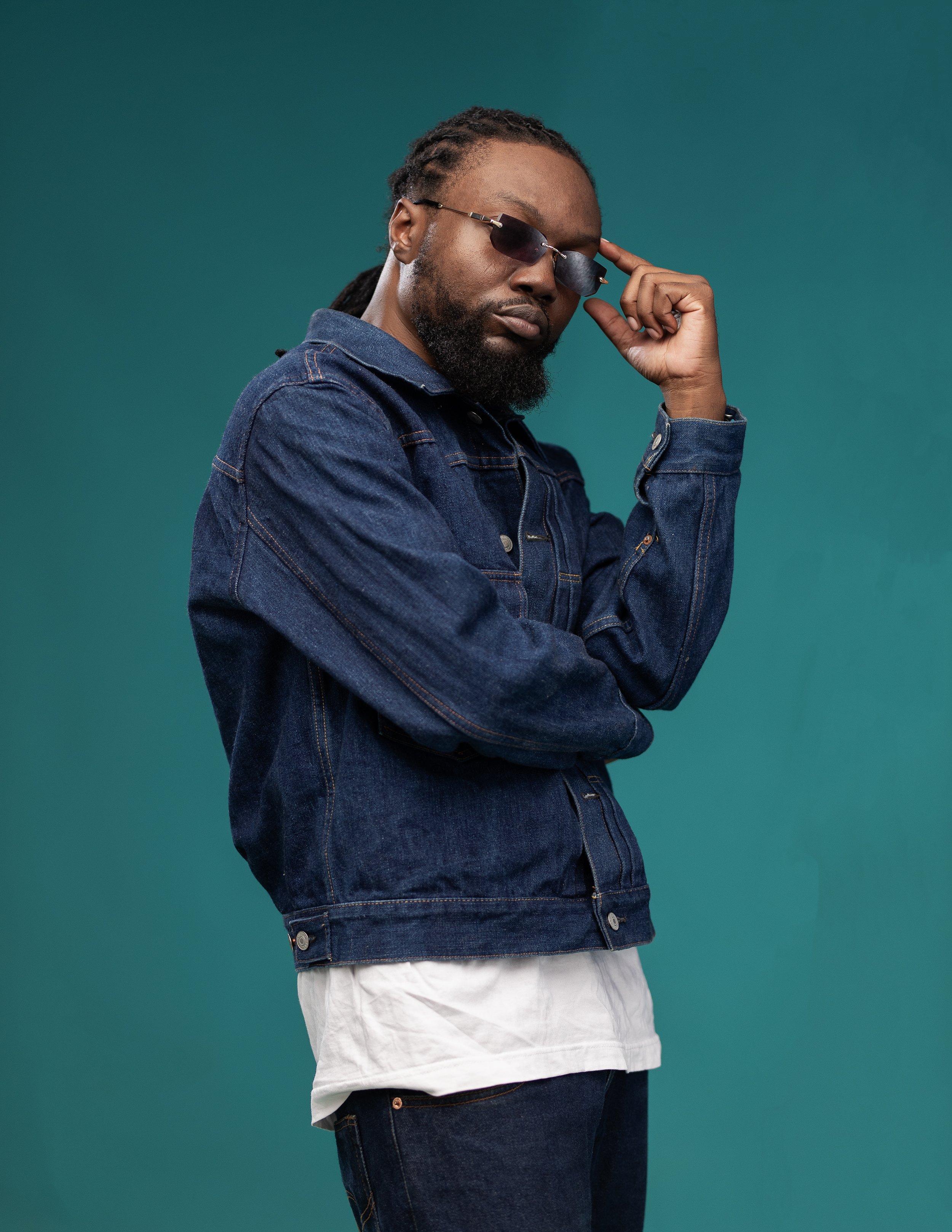 Person wearing sunglasses and a denim jacket against a teal background.