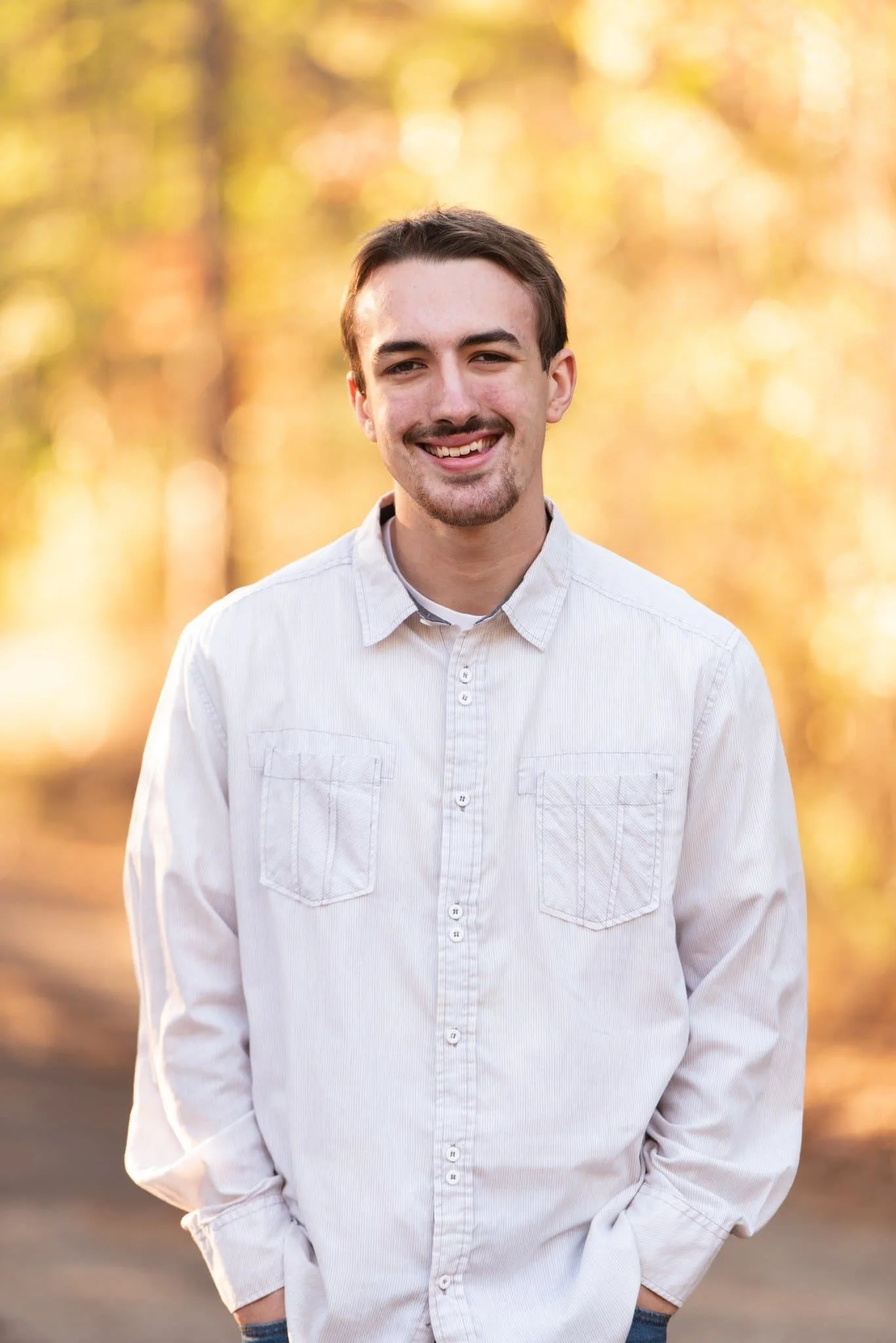 Professional outdoor portrait of a man in a light button‑up shirt and jeans standing in front of warm autumn trees.