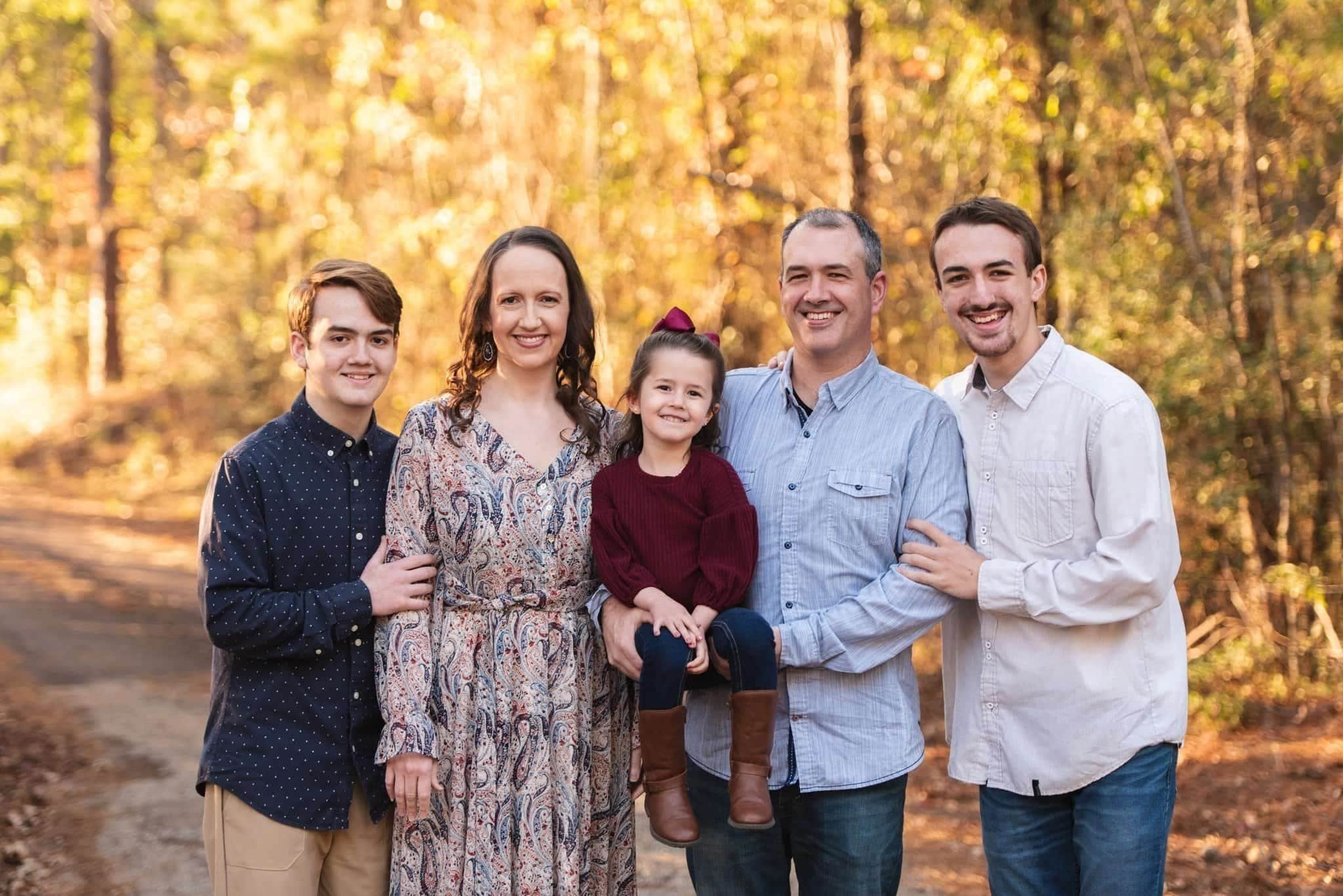 Family of five standing together outdoors in autumn, smiling in front of golden trees.