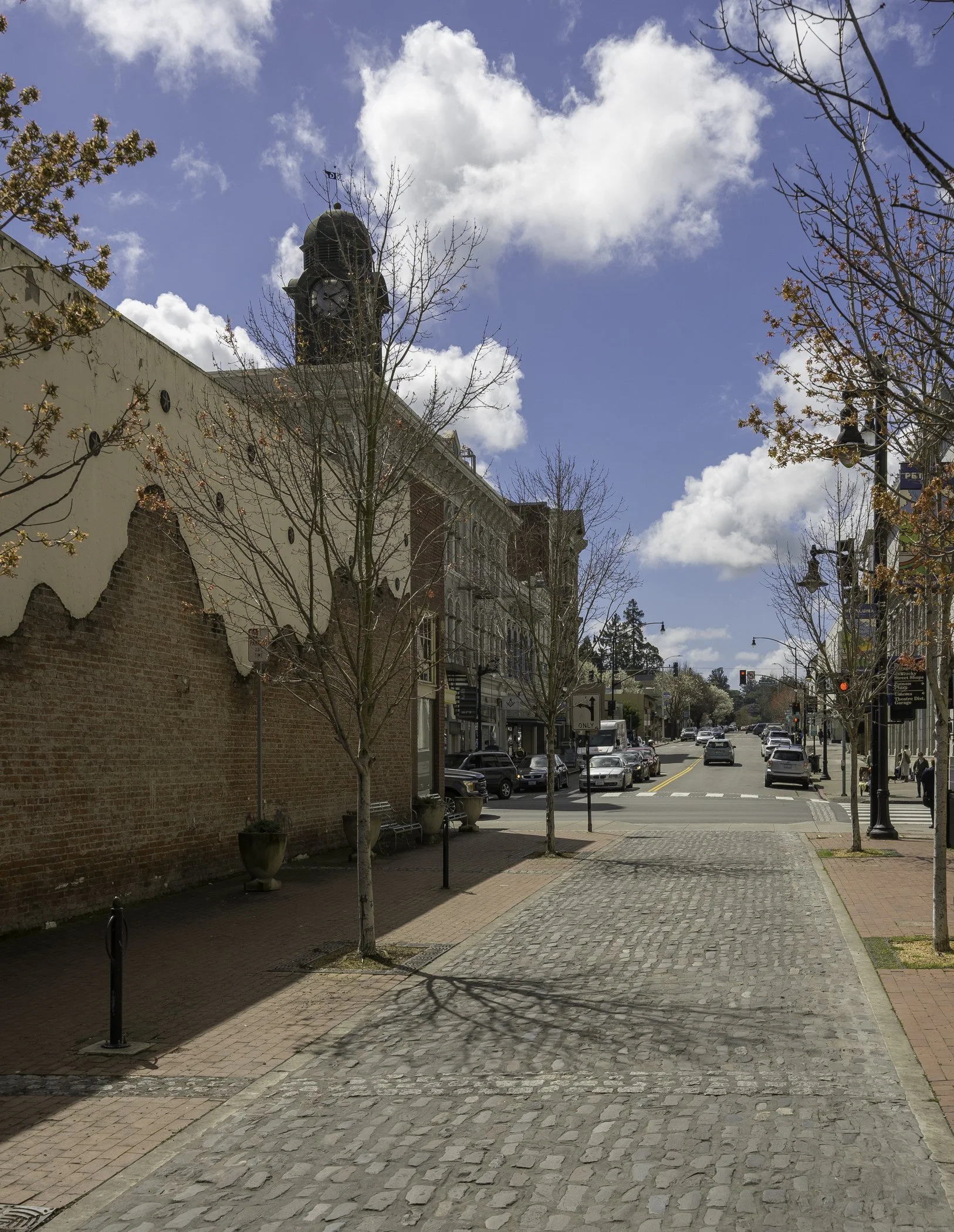 Cobblestone street with bare trees, brick buildings, and a distant clock tower under a partly cloudy sky.