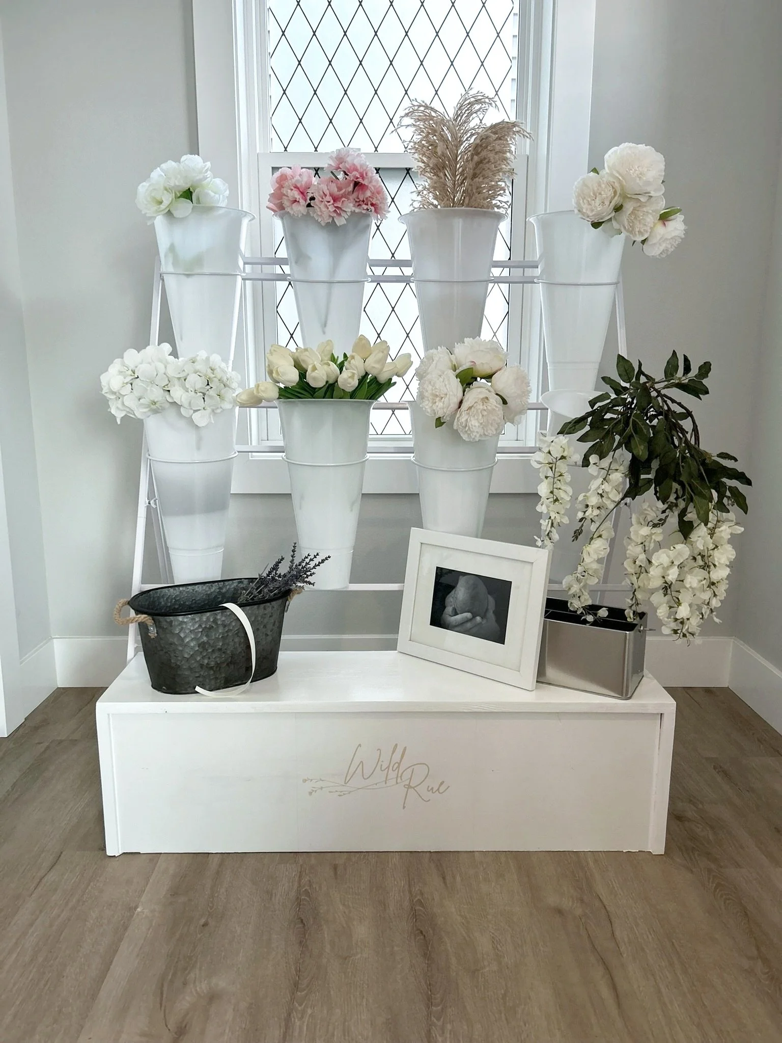 White display shelf with flower arrangements in white vases, black bucket with lavender, framed photo of a person, and a silver rectangular planter, set against a window with diamond-patterned grid.