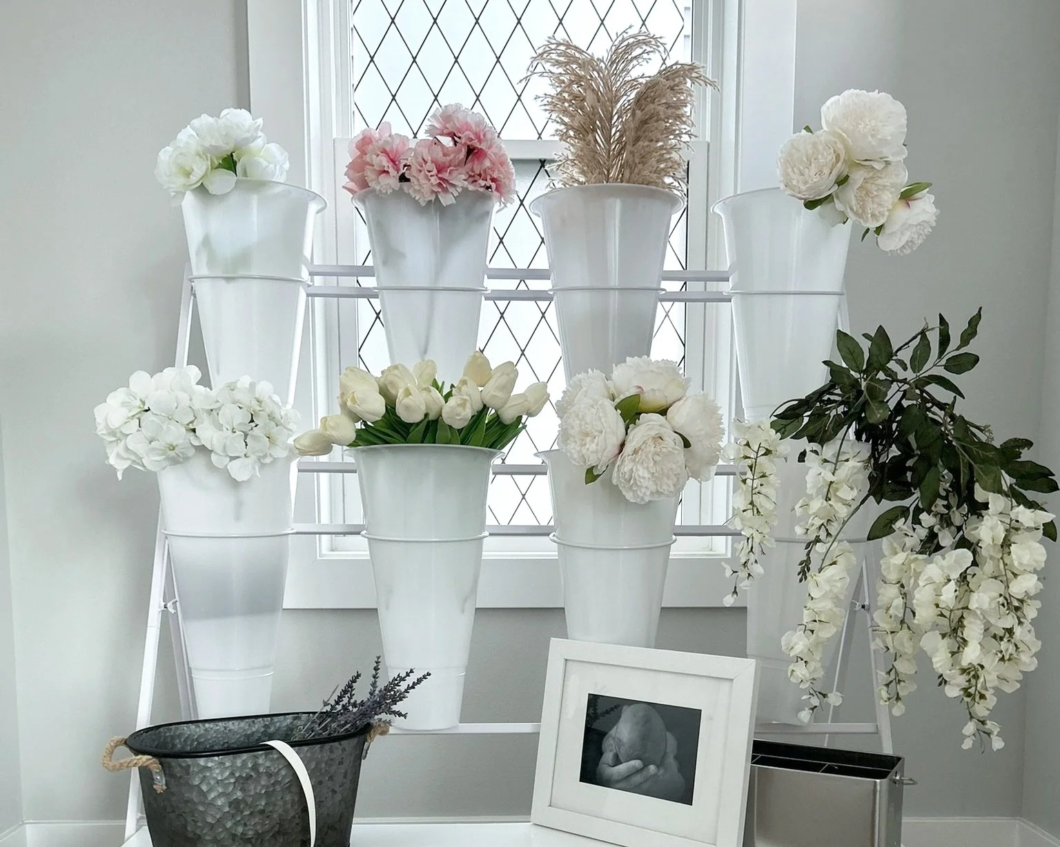 Display of white flowers in tall white vases on a white metal stand near a window with diamond-patterned glass, with a small framed black and white photo and a black basket in front.