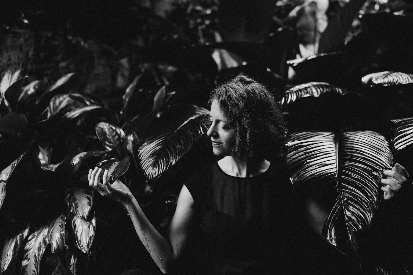 A woman with curly hair smiling and looking to her left, surrounded by large dark leaves, captured in black and white.