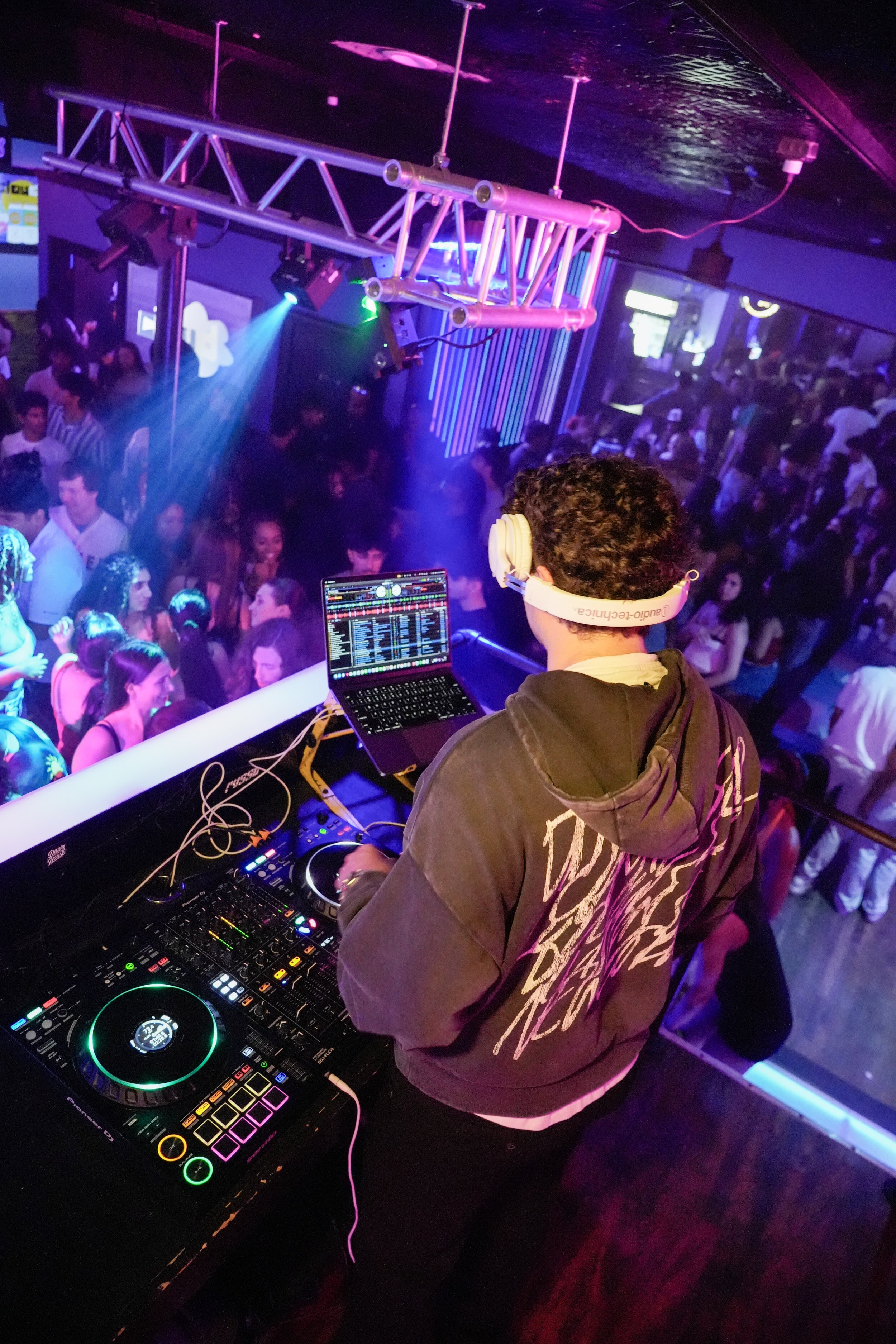 DJ with headphones standing at a mixing console in a crowded nightclub, with dancing people and colorful lights in the background.