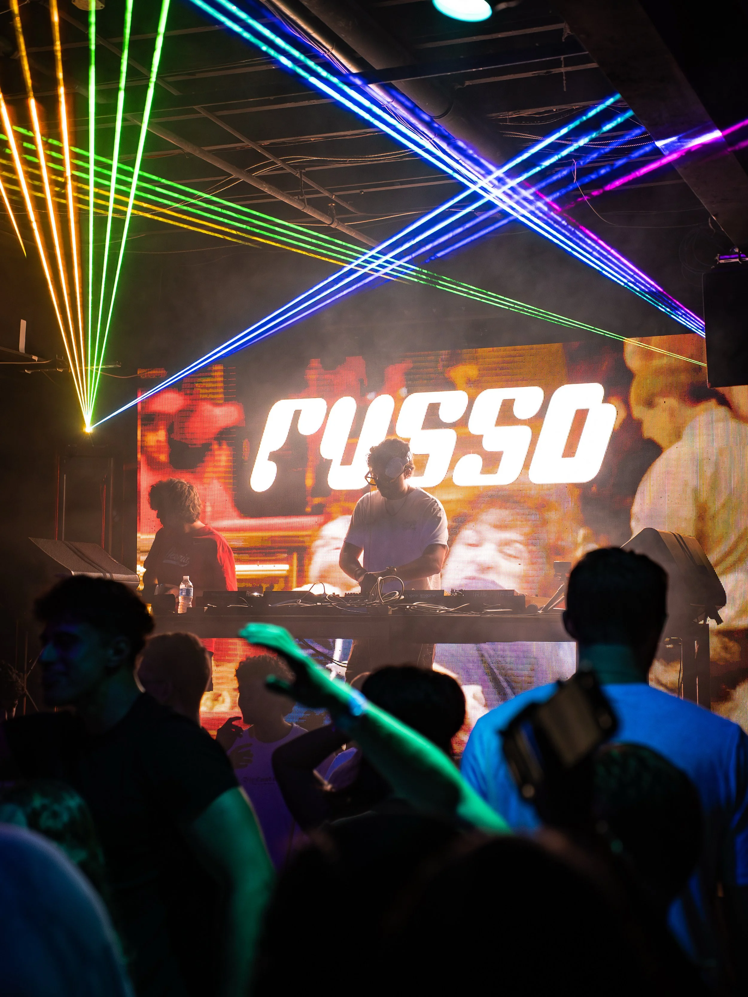 A DJ performing on stage at a nightclub with colorful laser lights and a large screen displaying the name 'FUSSO.' People in the crowd are dancing and enjoying the music.