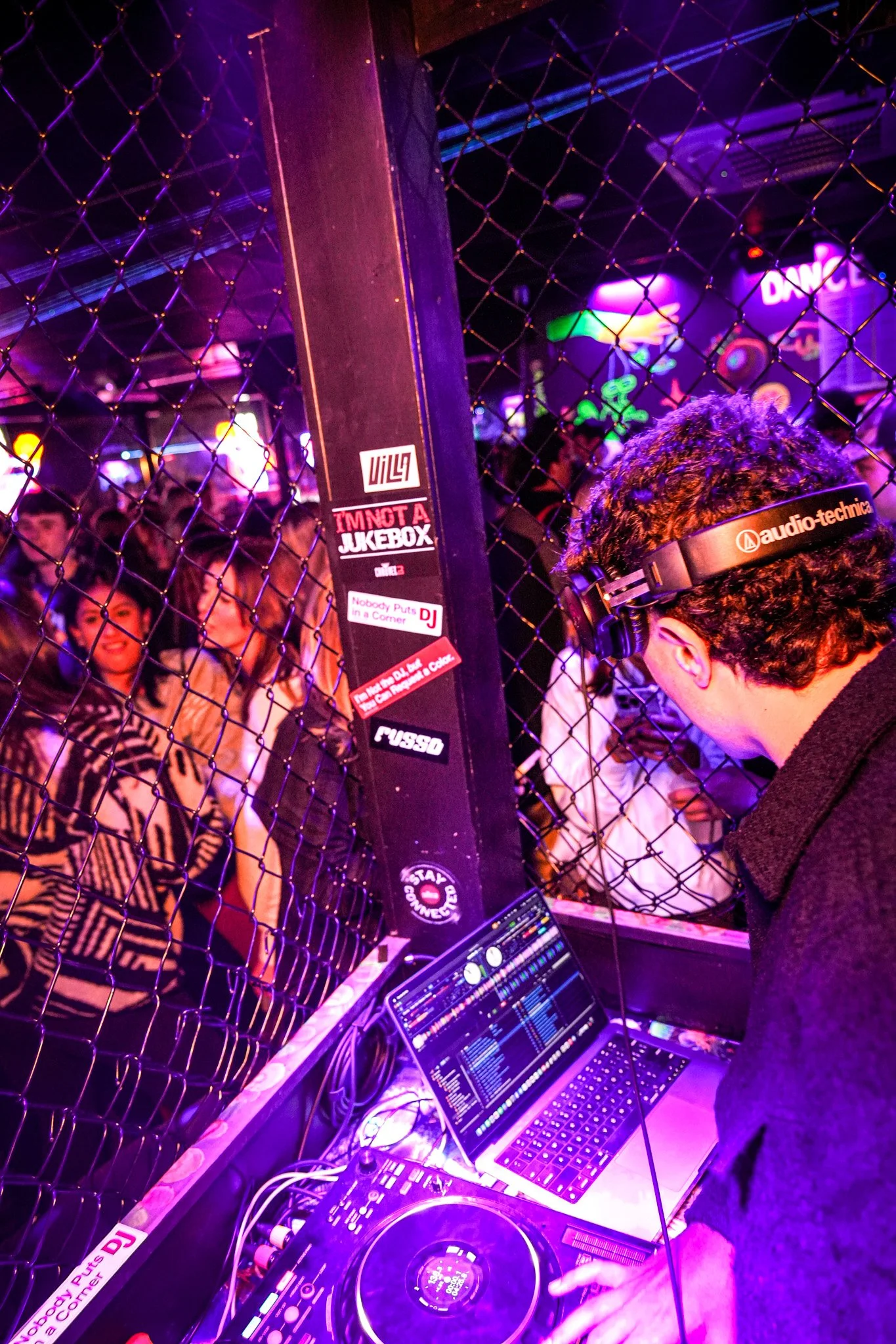DJ wearing headphones performing on a DJ mixer at a nightclub with a crowd dancing behind a wire fence.