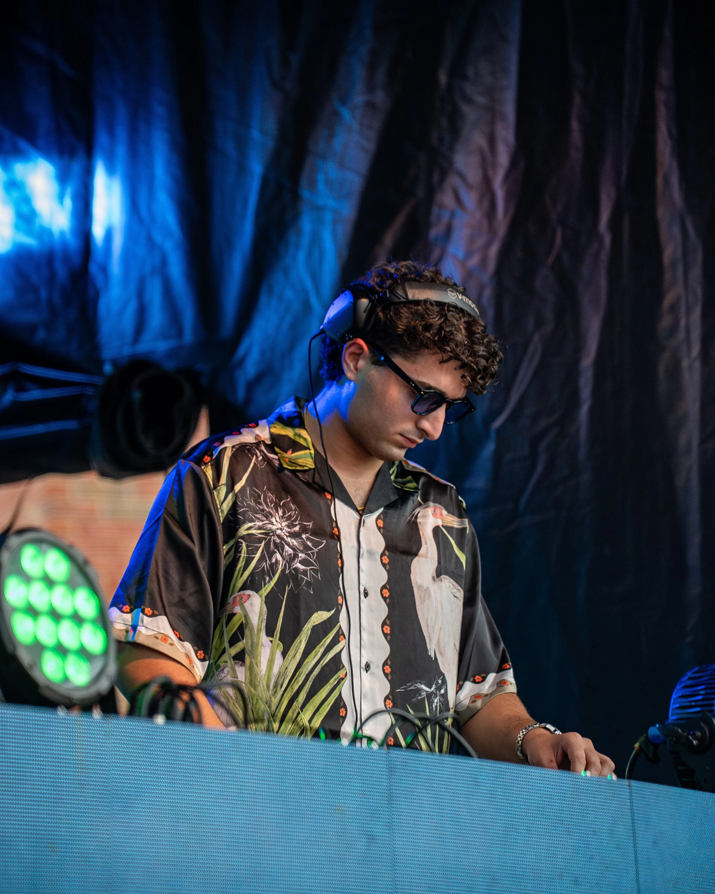 A young man with curly hair, wearing sunglasses, a Hawaiian shirt, and headphones, is DJing at a music event behind a blue-colored DJ booth with stage lights around him.