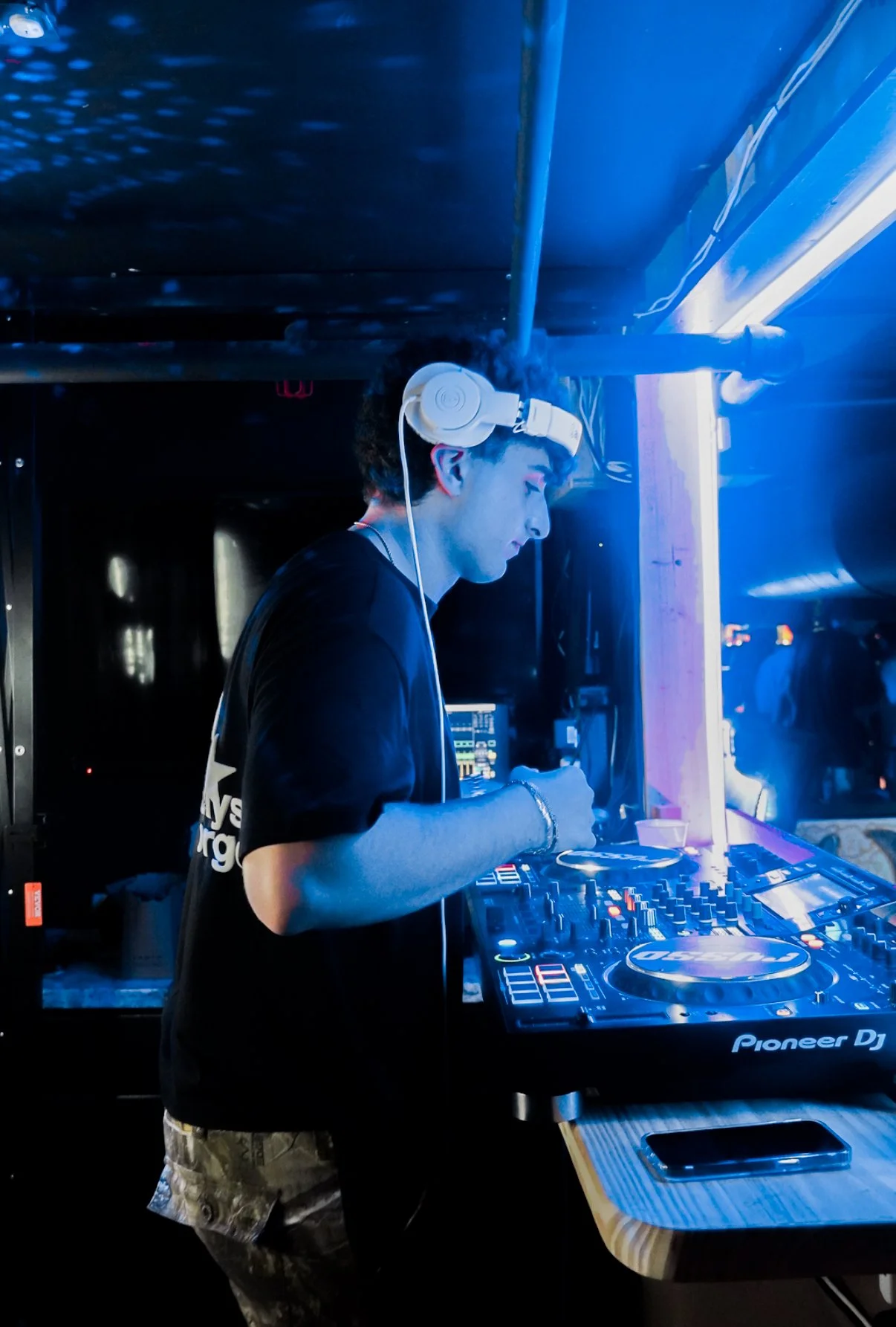 A young male DJ wearing headphones, mixing music on a Pioneer DJ controller at a nightclub or party with blue lighting.