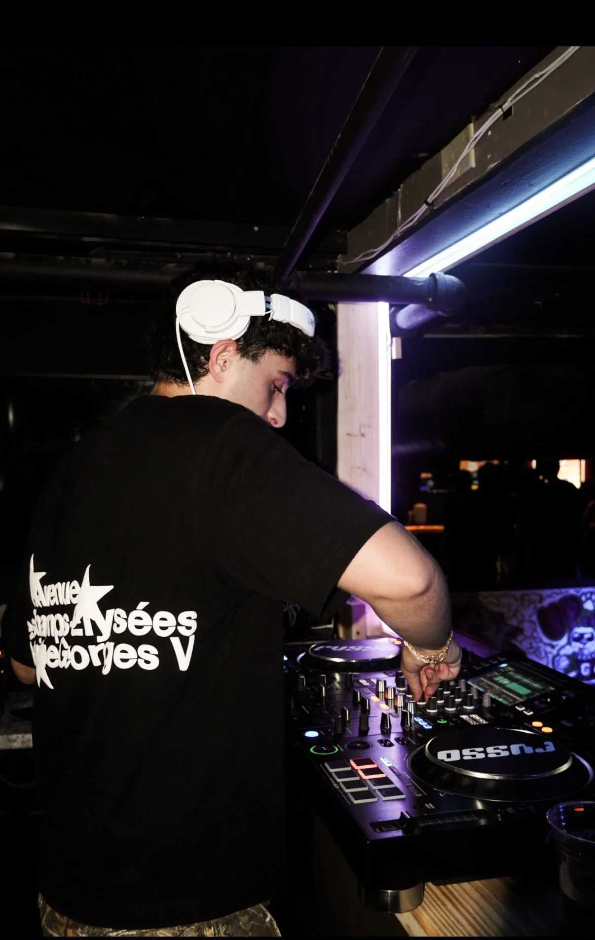 A young male DJ wearing white headphones, a black t-shirt, and camouflage pants, mixing music at a DJ console in a dimly lit club or bar with purple lighting.