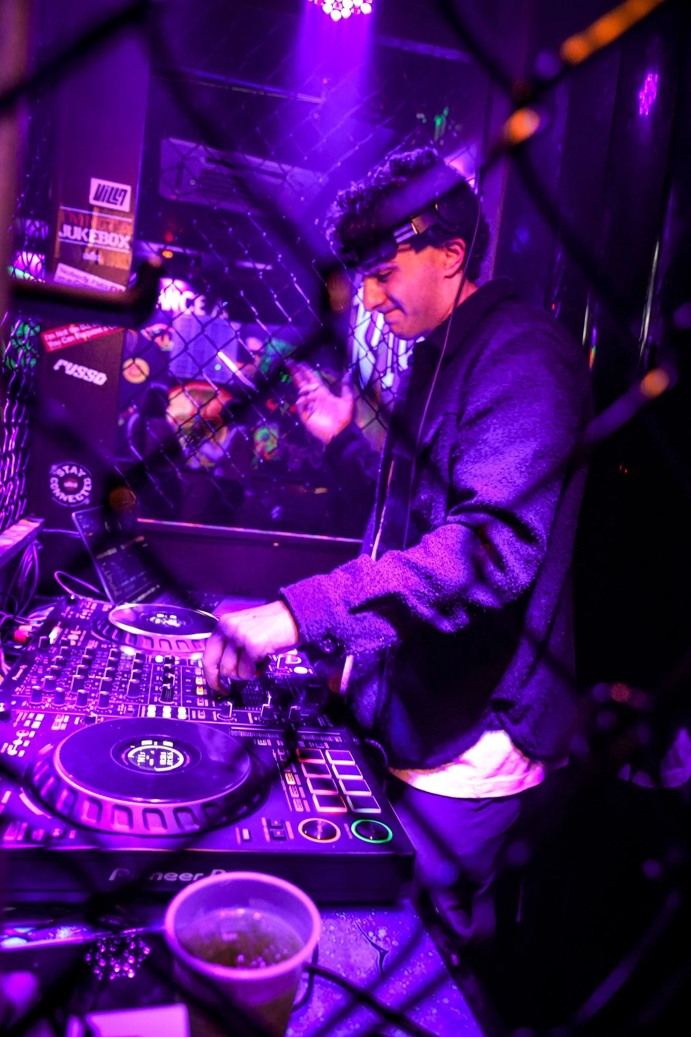 A DJ mixes music in a club with purple lighting, wearing headphones and a black hoodie, surrounded by DJ equipment and a chain-link fence.