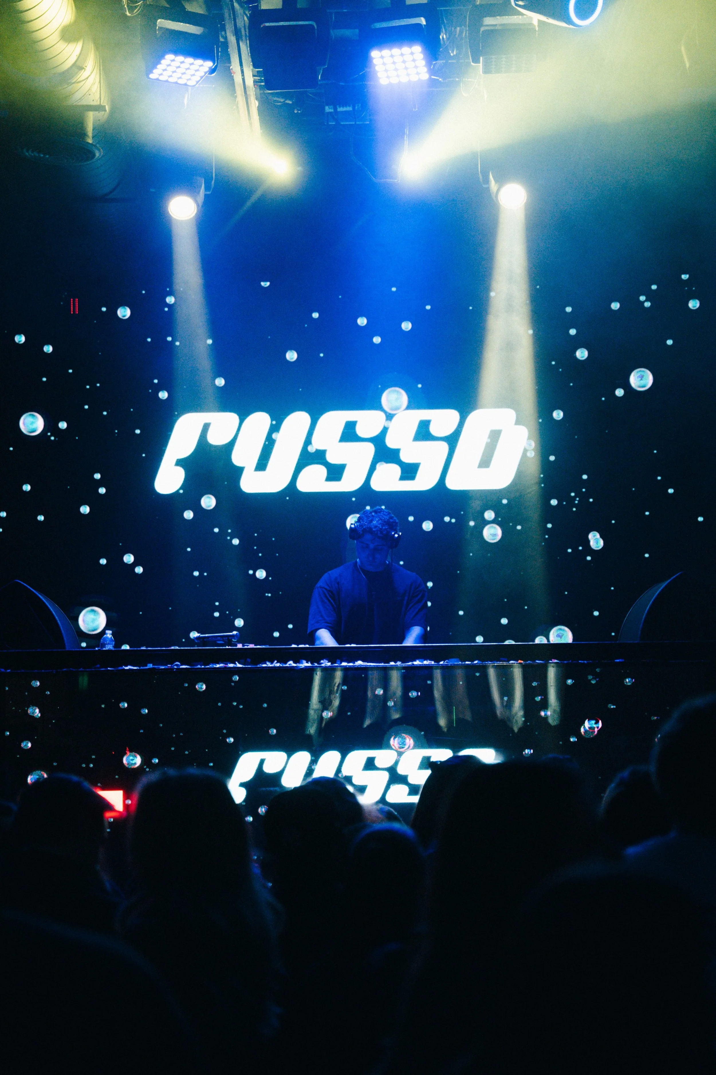 DJ performing on stage with a digital display behind reading 'RUSSO' and audience in foreground.