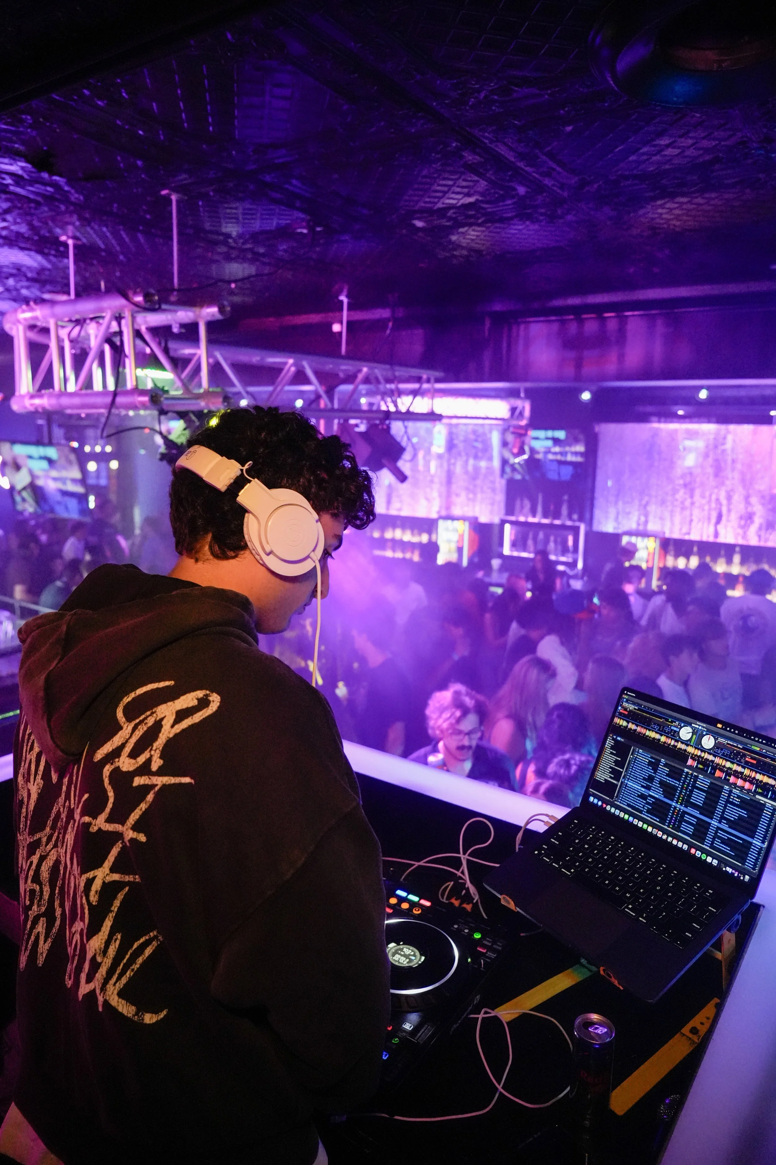 DJ wearing headphones and operating DJ equipment at a nightclub with purple lighting and a crowded dance floor.
