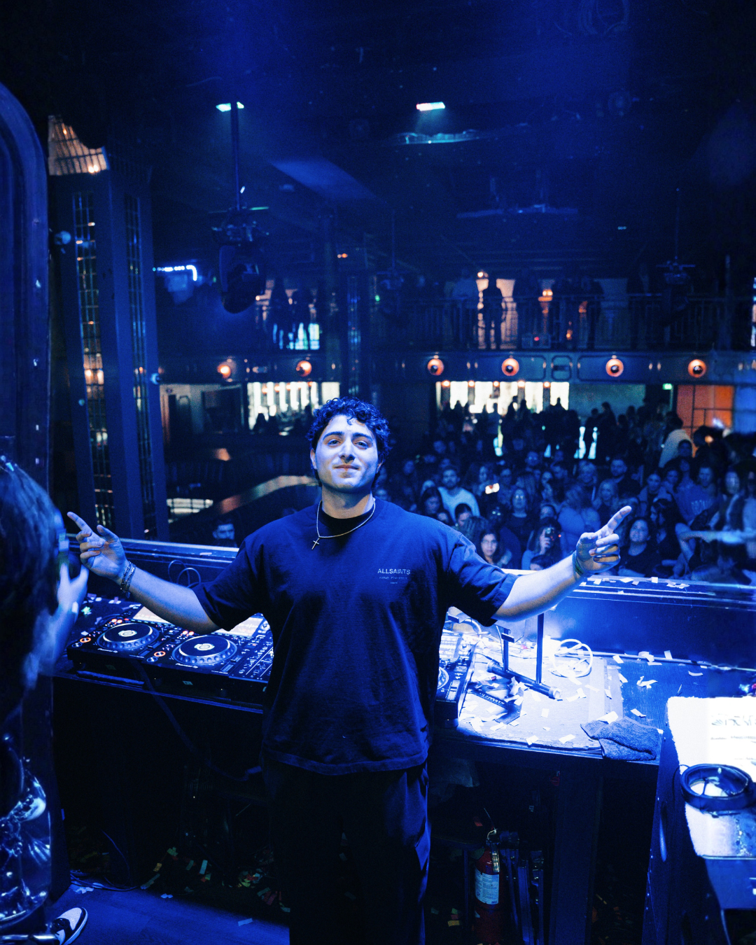 A DJ with curly hair and a black t-shirt smiling and pointing with both hands behind a DJ booth at a nightclub with a crowd dancing in the background, blue lighting, and a balcony.