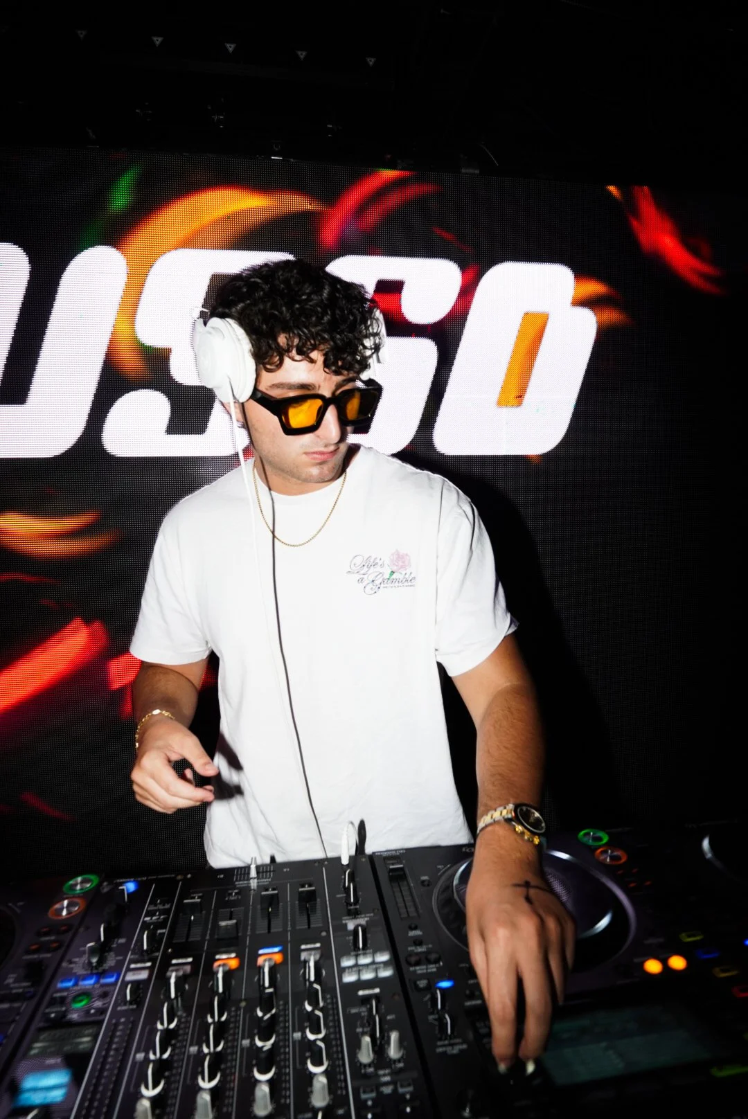 A male DJ with curly black hair, wearing yellow sunglasses, white t-shirt, headphones, and jewelry, operating a DJ mixer in front of a large screen displaying the number '150' with colorful abstract graphics.