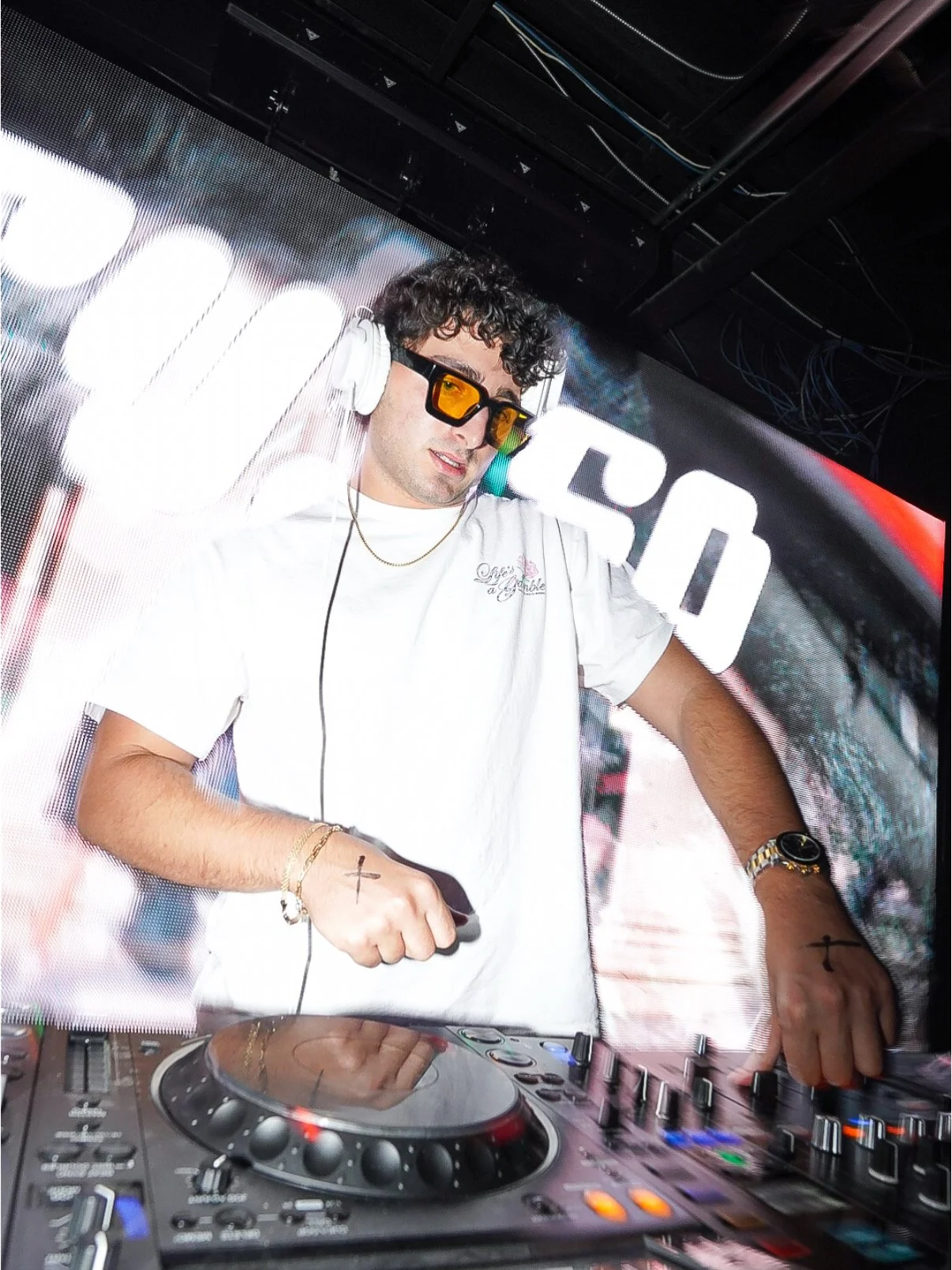 A DJ with curly hair wearing sunglasses and headphones, using a DJ mixer in a club with a digital display behind him.