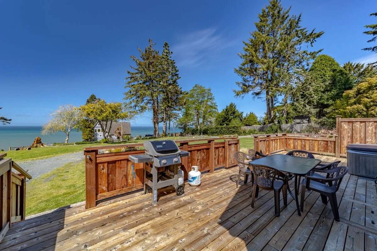  wooden deck with a table and chairs, barbecue grill, scattered trees, green lawn, fence, house, and ocean view in the background under a clear blue sky.