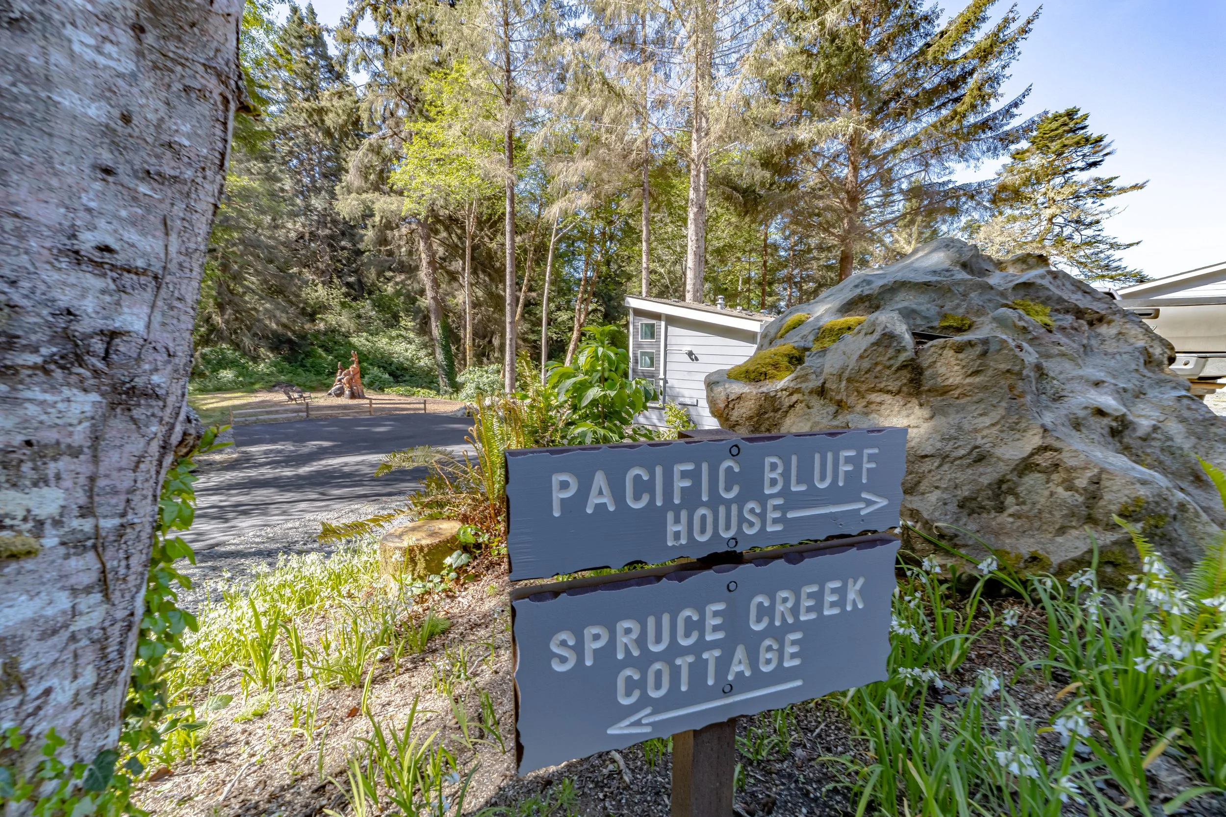 A sign indicating directions to Pacific Bluff House and Spruce Creek Cottage, situated near a large moss-covered rock and surrounded by greenery and tall trees.