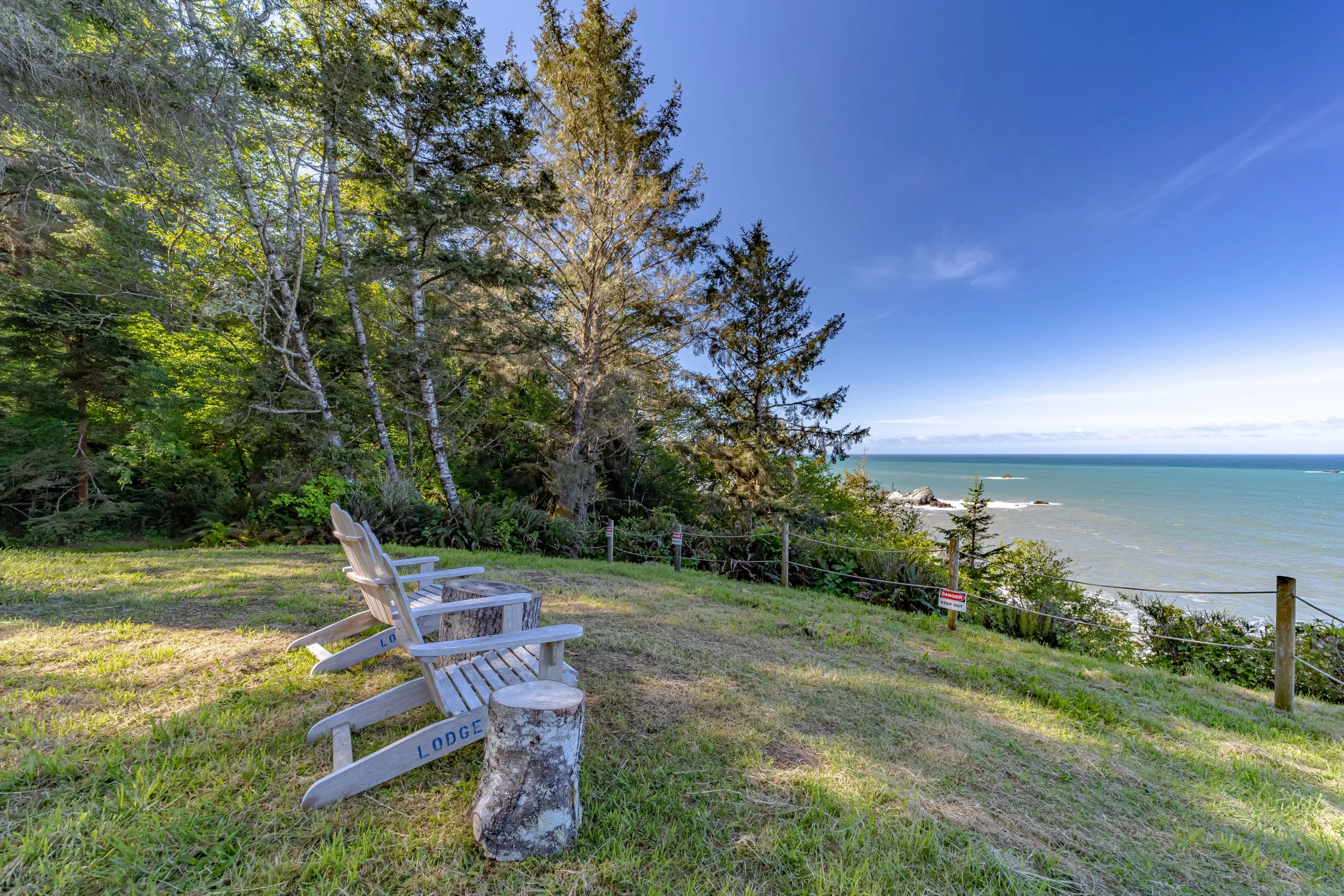 Three wooden Adirondack chairs facing the ocean on a grassy hill, surrounded by trees, with a clear blue sky overhead.