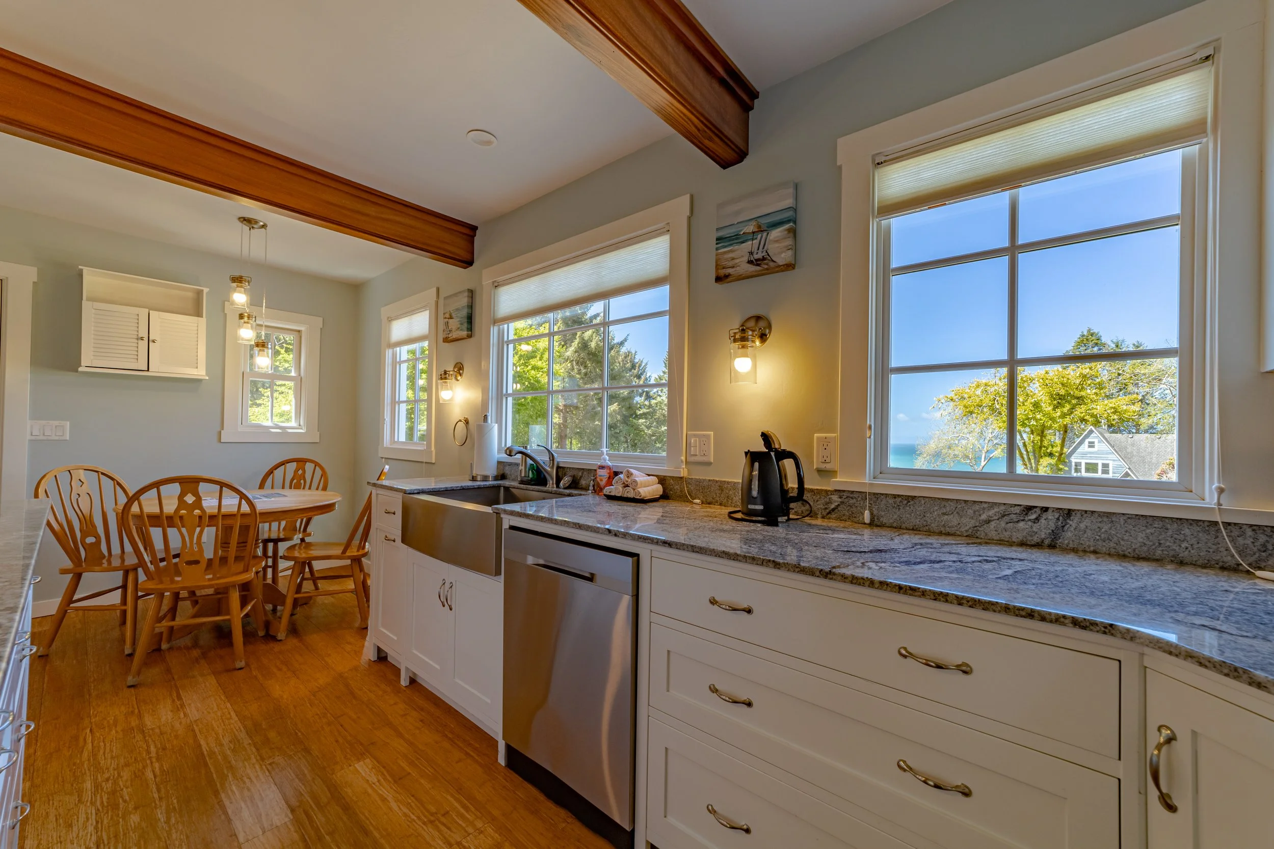 Kitchen with white cabinets, granite countertops, stainless steel dishwasher, black electric kettle, and large windows showing trees and blue sky.