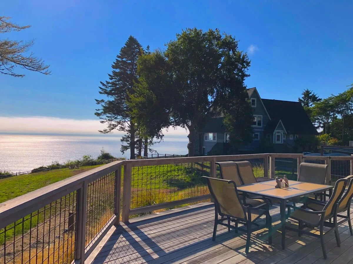 A wooden deck with a table and chairs overlooking a shore with water, trees, and a house in the distance under a clear blue sky.