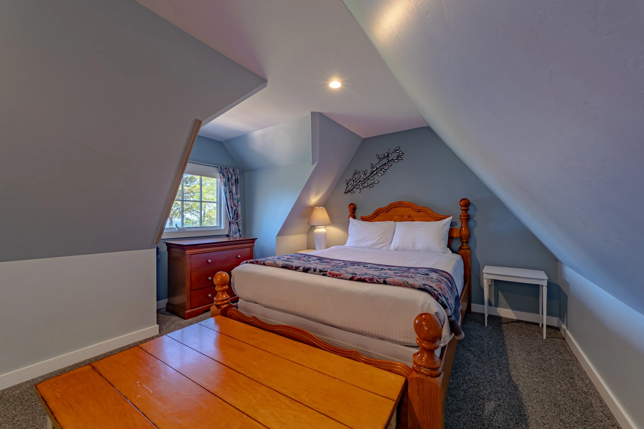 Bedroom with a wooden bed, a nightstand with a lamp, a dresser near a window, and a wooden table at the foot of the bed in an attic space with sloped ceilings and blue walls.