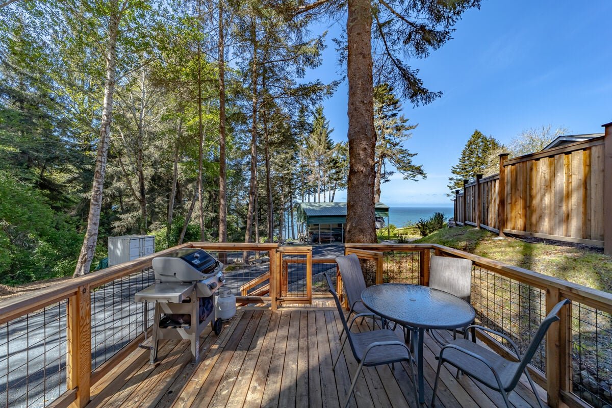 A wooden deck with a barbecue grill and a round glass table with four chairs, surrounded by trees and overlooking the ocean.